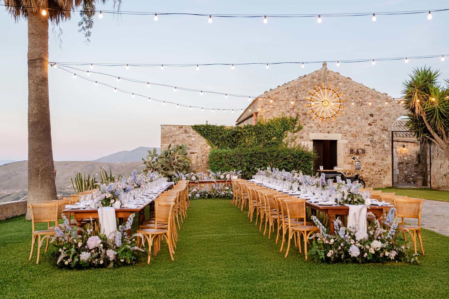 Romantic outdoor wedding reception setup with long floral tables, string lights, and a historic stone chapel backdrop.