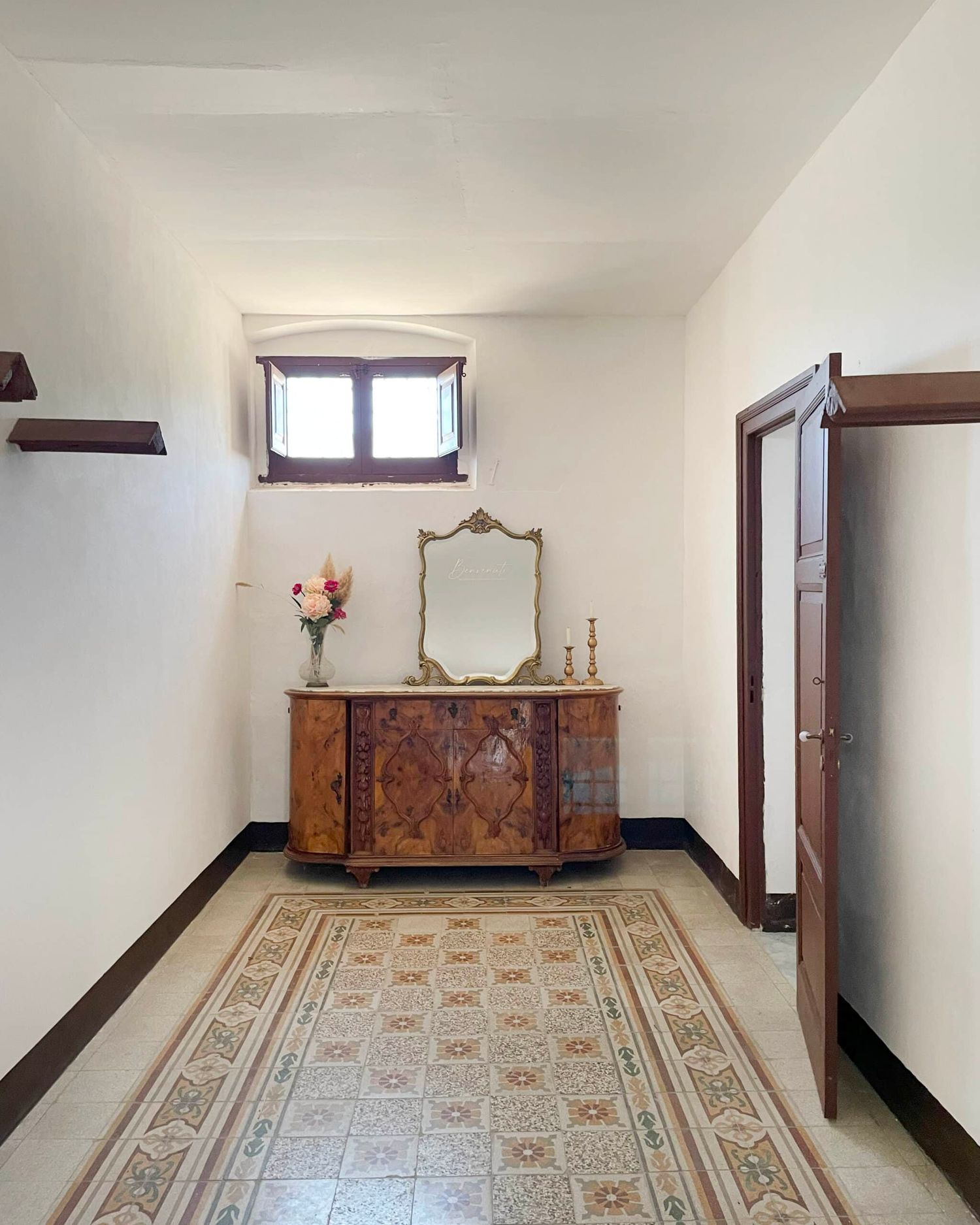 Elegant hallway with ornate wooden dresser, brass mirror, floral tile floor, and decorative vase with flowers.