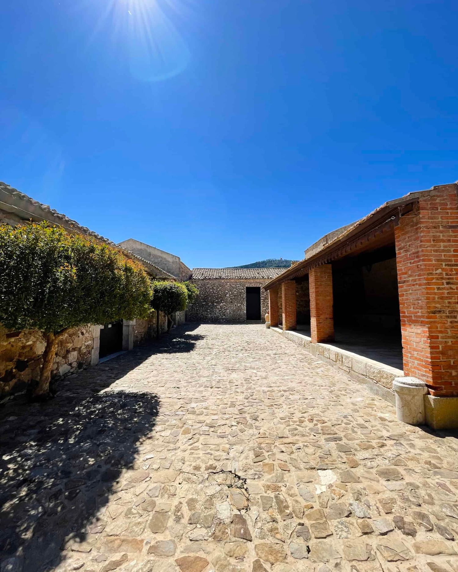 Rustic cobblestone courtyard with brick and stone buildings under bright sunny blue sky.