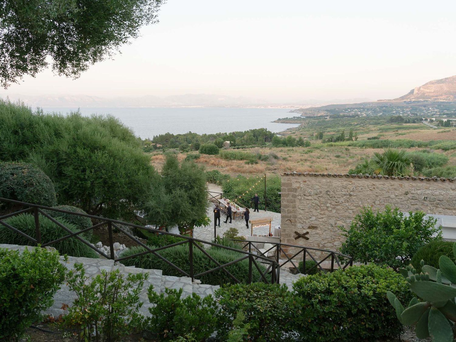 Scenic Mediterranean coastal view from stone terrace with lush greenery, stairs, and calm sea in the background at dusk. Scopello, Sicily.