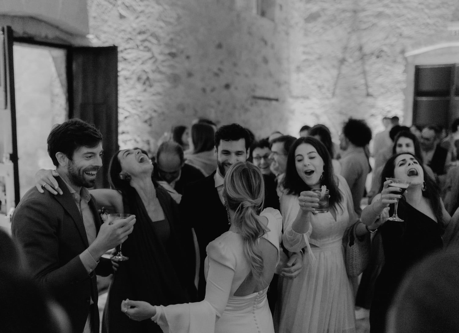 Black and white photo of wedding guests laughing and celebrating with drinks at a lively rustic stone venue reception.