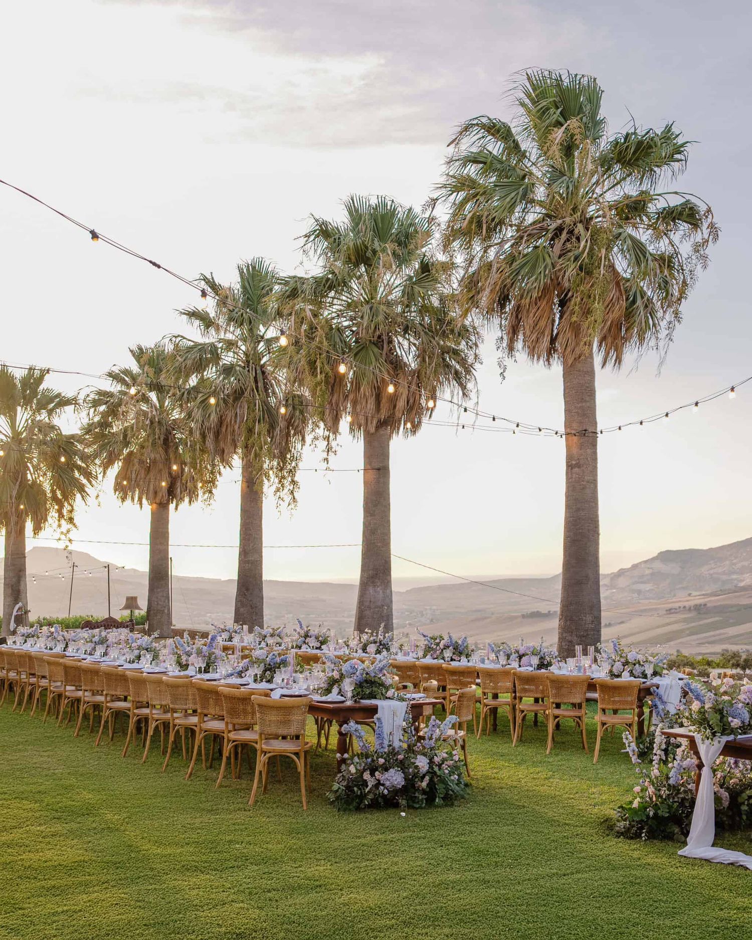 Elegant outdoor wedding reception with long tables, wooden chairs, string lights, and tall palm trees at sunset.