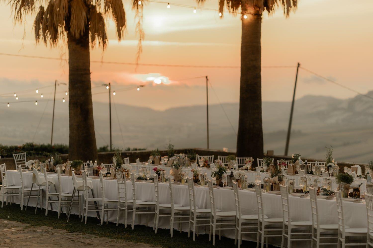Outdoor wedding reception with long white tables set at sunset, framed by palm trees and scenic mountain views.