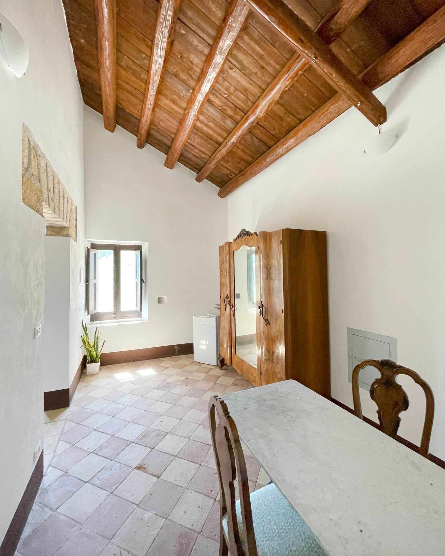 Spacious rustic room with high wooden beam ceiling, terracotta tile floor, antique wardrobe, and dining table.