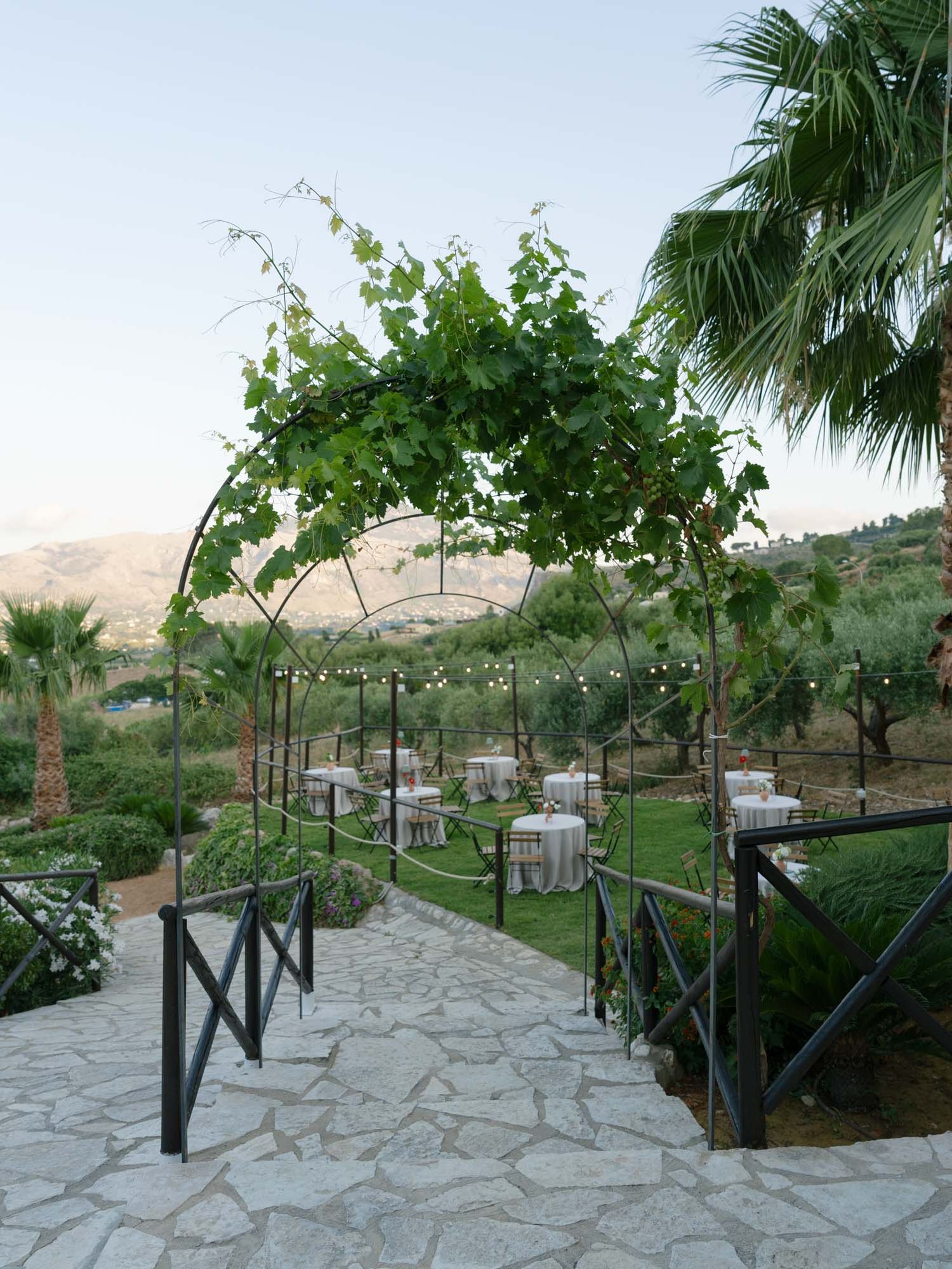 Stone pathway leads through a vine-covered arch to an elegant outdoor dining area with mountain views. Mulino 1220 in Scopello.