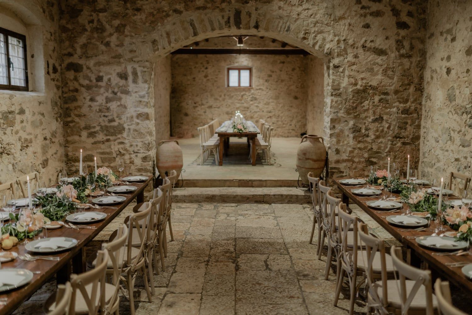 Elegant rustic wedding dinner setup with long wooden tables, candles, and floral decor inside a stone-arched venue.