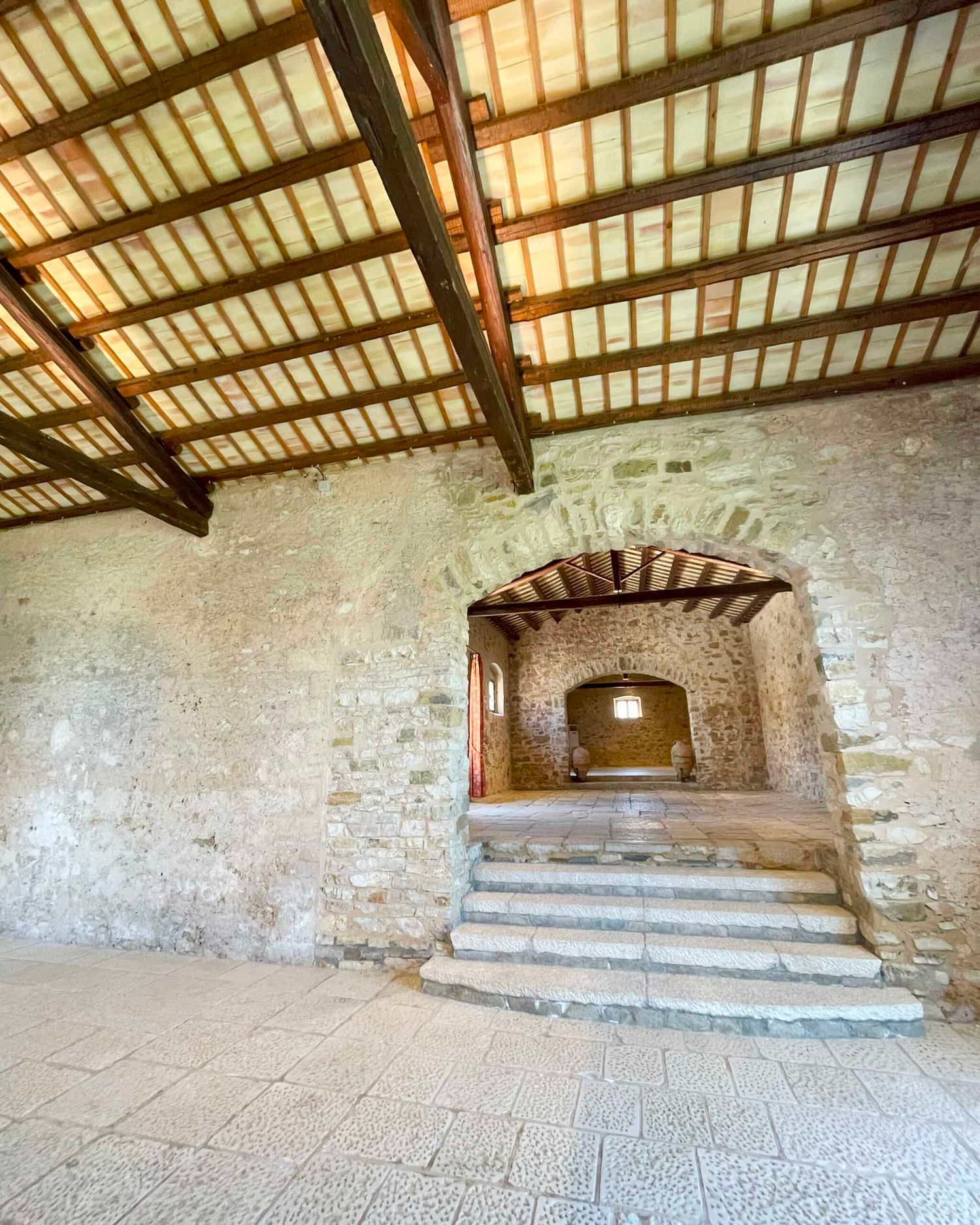 Historic stone building interior featuring a vaulted wooden ceiling, arched alcove, and stone steps leading to a doorway.