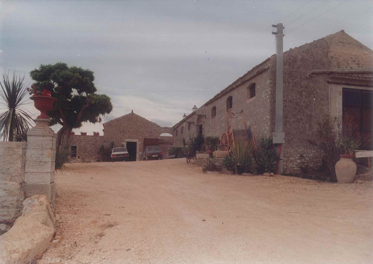 Rural stone farmhouse complex with dirt road, large terracotta urns, and desert plants.