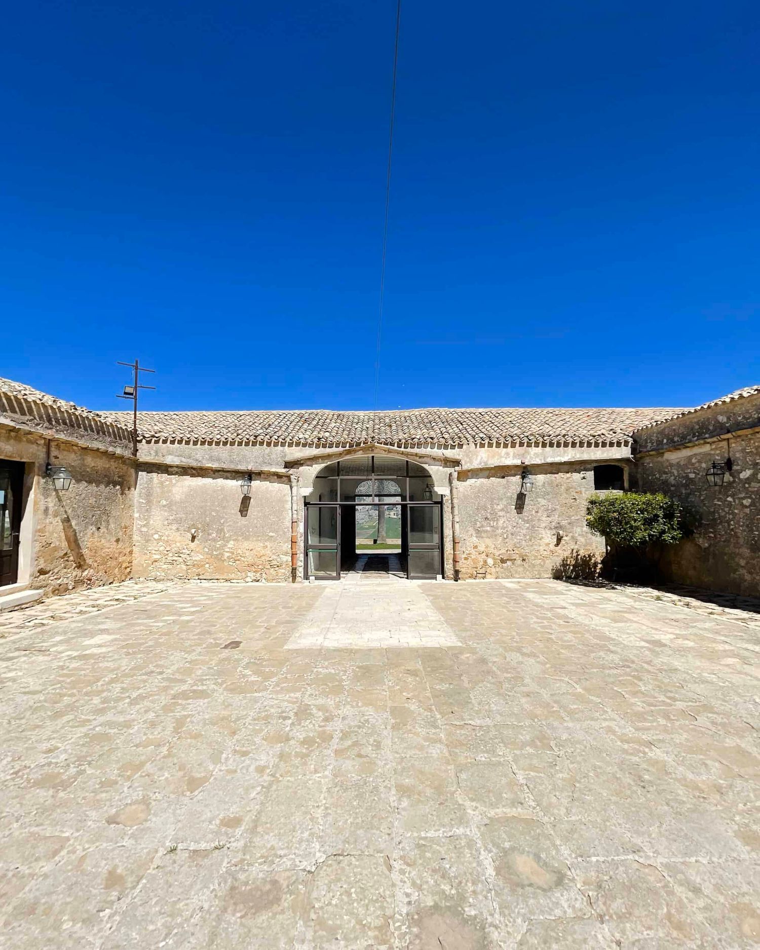 Restored stone building facade with arched entrance and large paved courtyard under blue sky.
