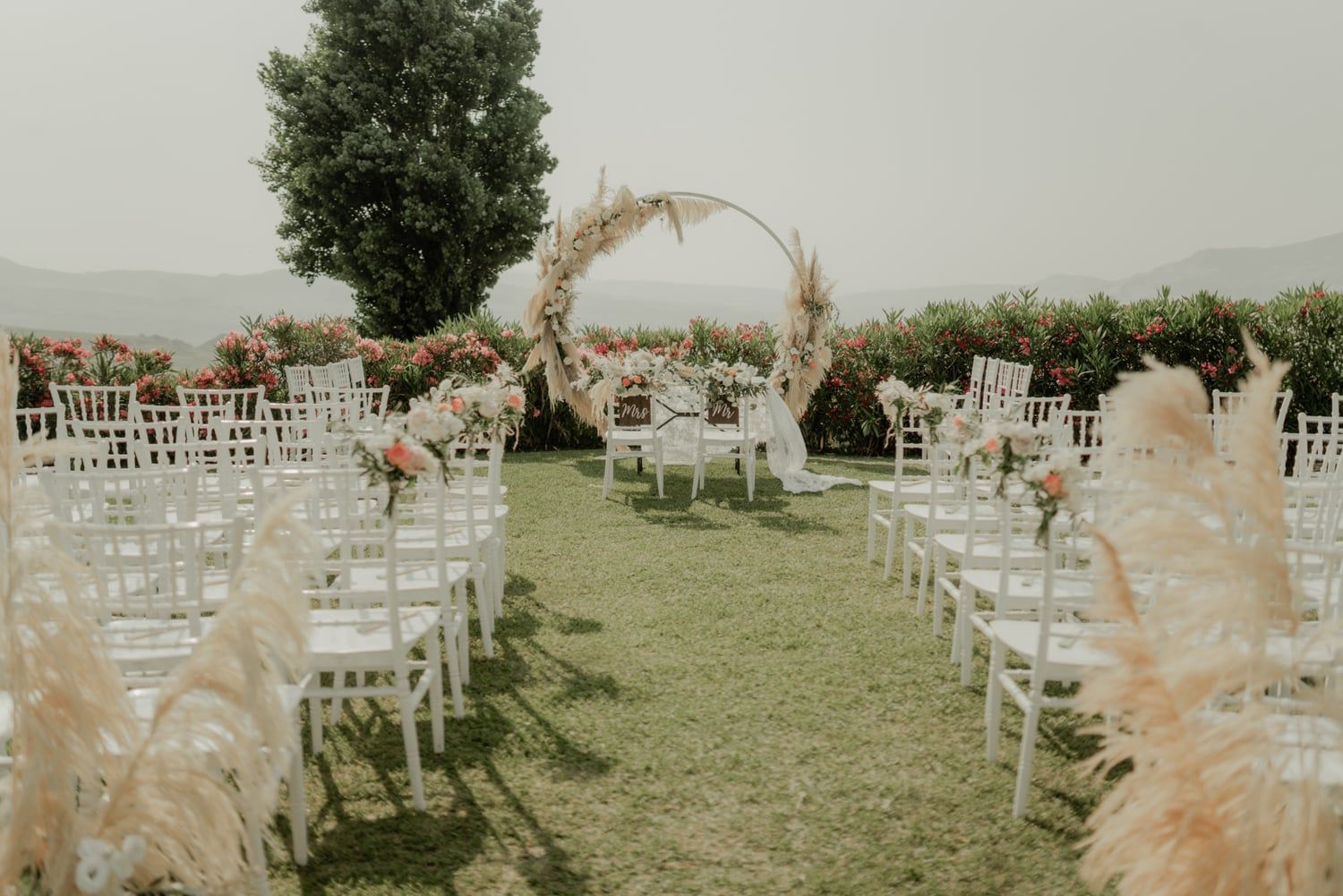 Outdoor wedding ceremony setup with white chiavari chairs, pampas grass, and a floral circular arch.