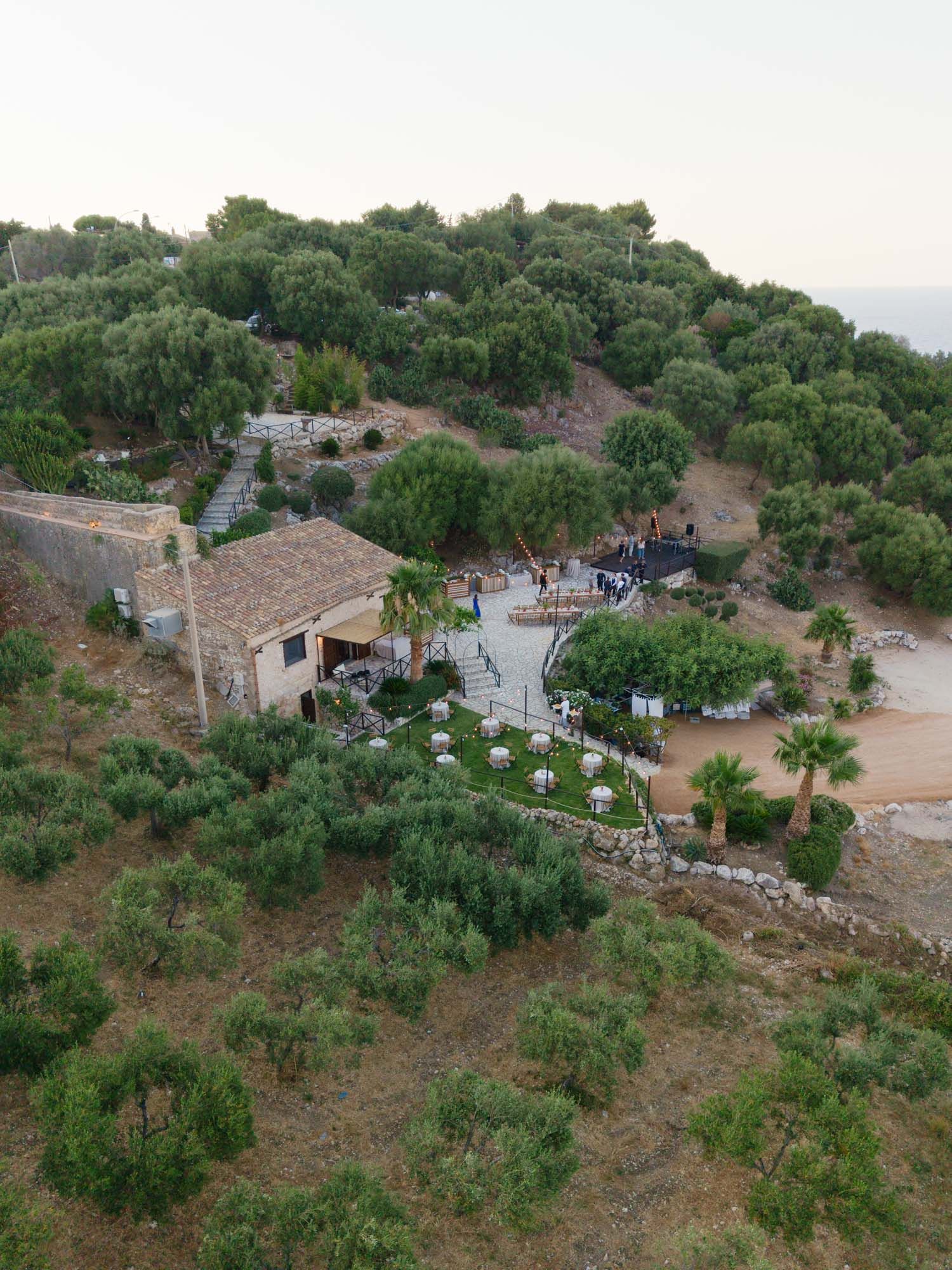 Aerial view of a rustic stone villa surrounded by olive trees and palm trees on a Mediterranean hillside. Mulino 1220 in Scopello.