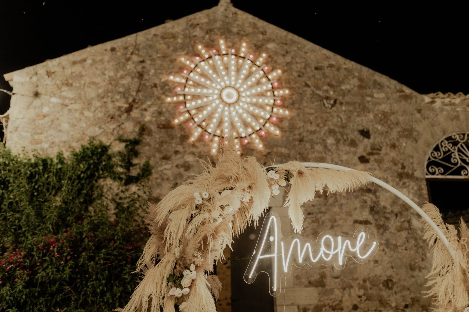 Romantic outdoor wedding venue with neon Amore sign, pampas grass arch, and glowing rose window at night.