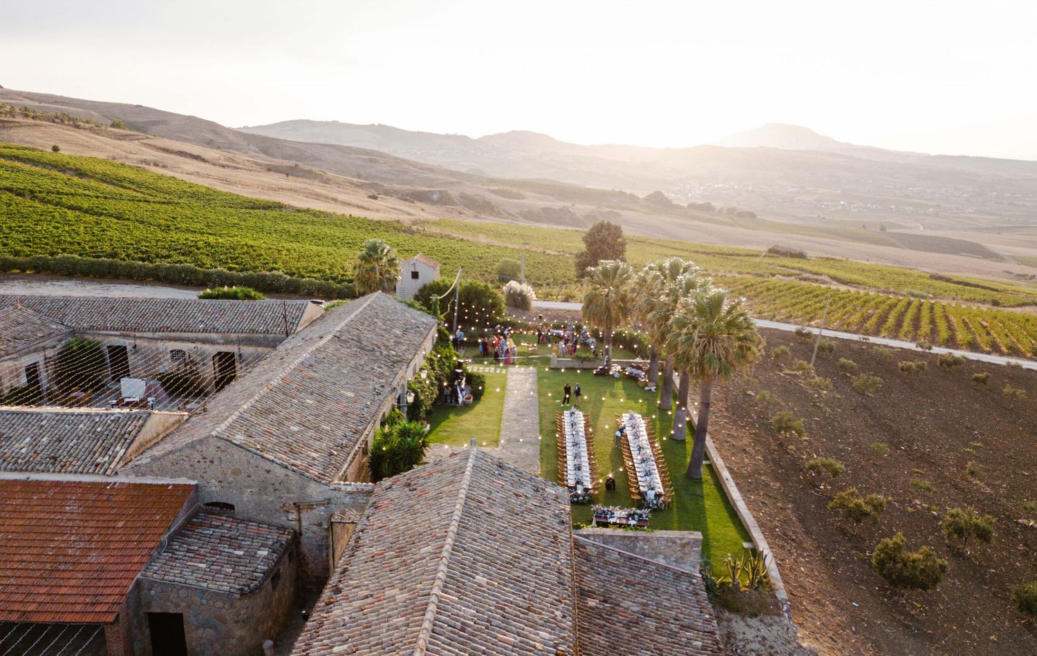 Aerial view of rustic vineyard wedding venue surrounded by rolling hills and lush grapevines at sunset.