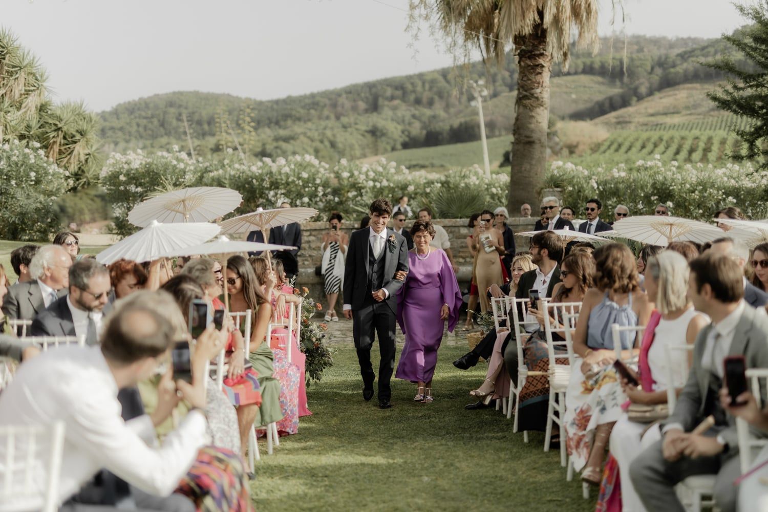Outdoor wedding ceremony on lush green lawn with guests seated in white chairs, surrounded by rolling hills and palm trees.