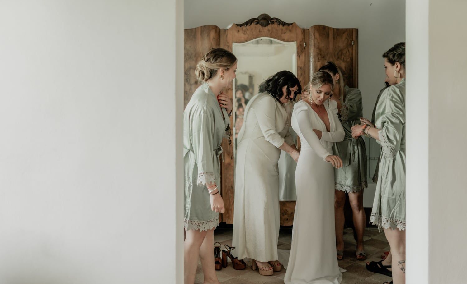 Bride being helped into her wedding dress by bridesmaids wearing sage green robes in a bright dressing room.