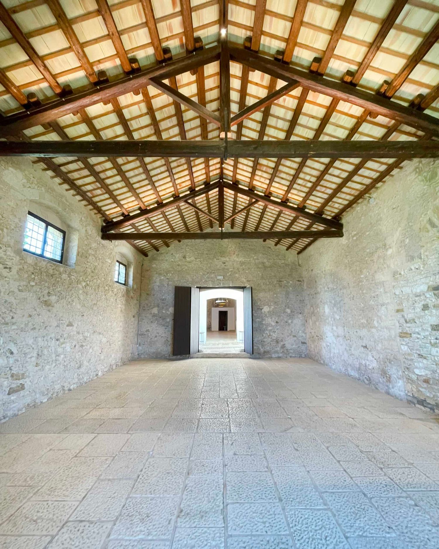 Large renovated stone barn with vaulted wooden beam ceiling, stone tile floor, and arched doorway leading to courtyard.