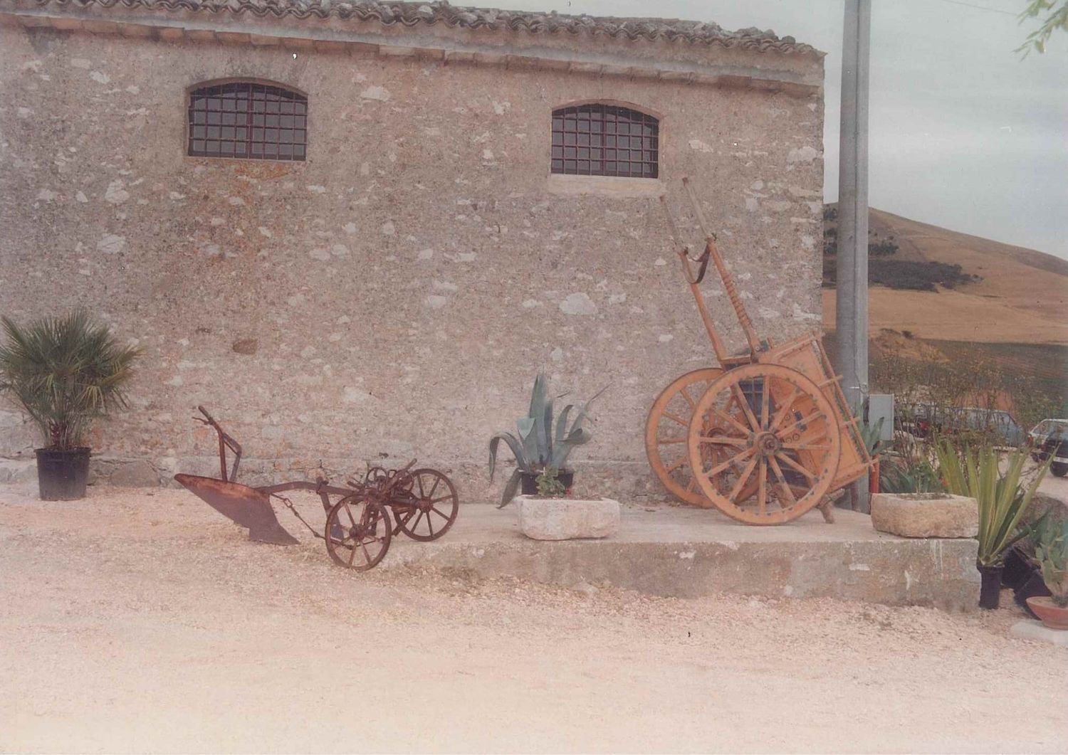 Rustic stone farmhouse exterior with antique wooden cart and vintage plow displayed on gravel courtyard.