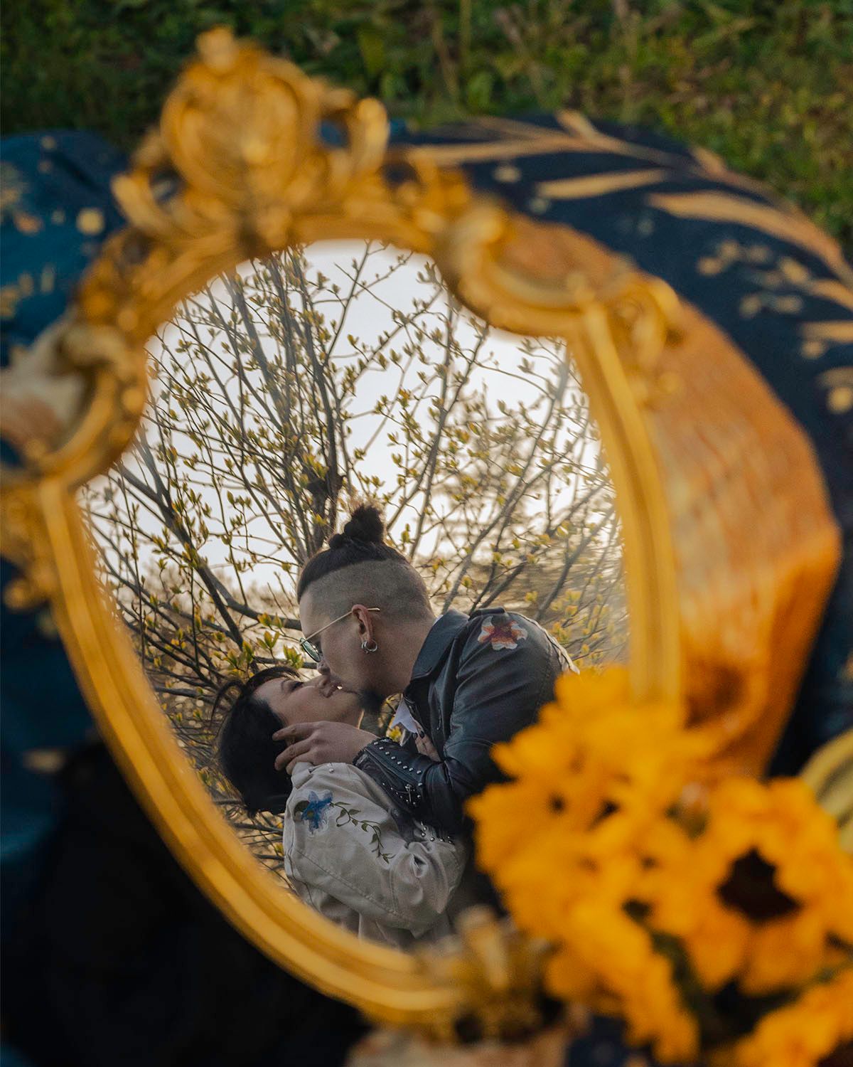 Couple kissing reflected in ornate gold mirror surrounded by sunflowers and spring trees outdoors.