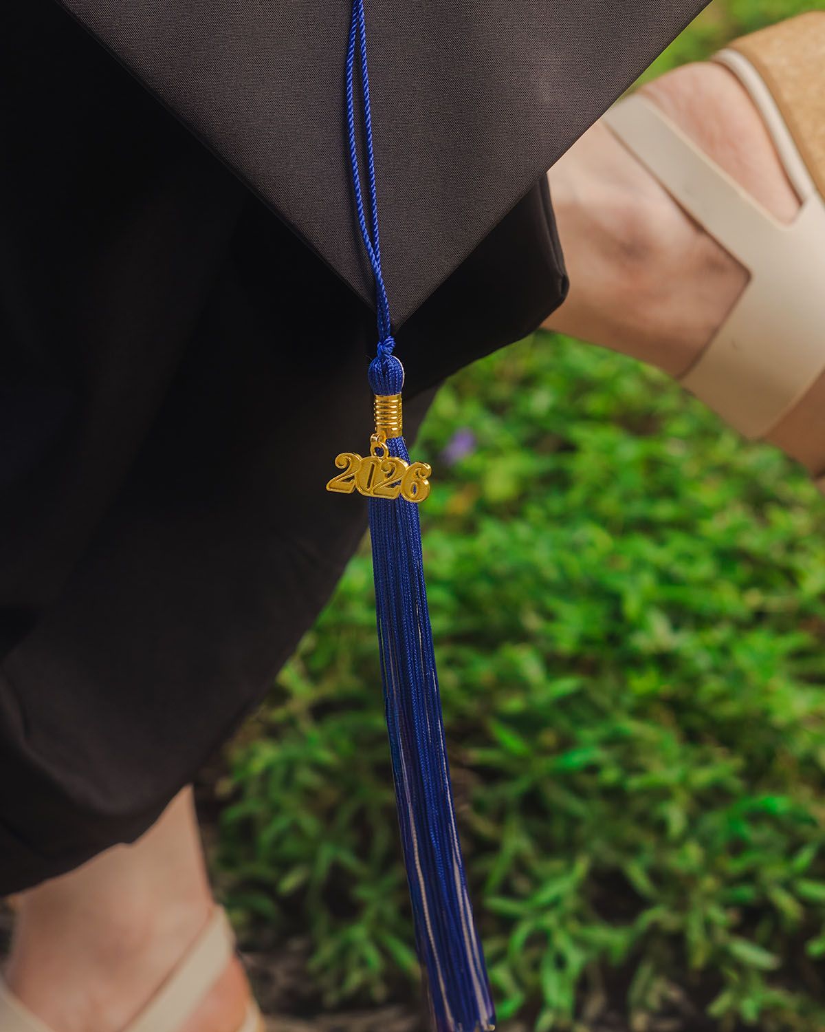 Close-up of a blue graduation tassel with a gold 2026 charm on a black academic gown outdoors.