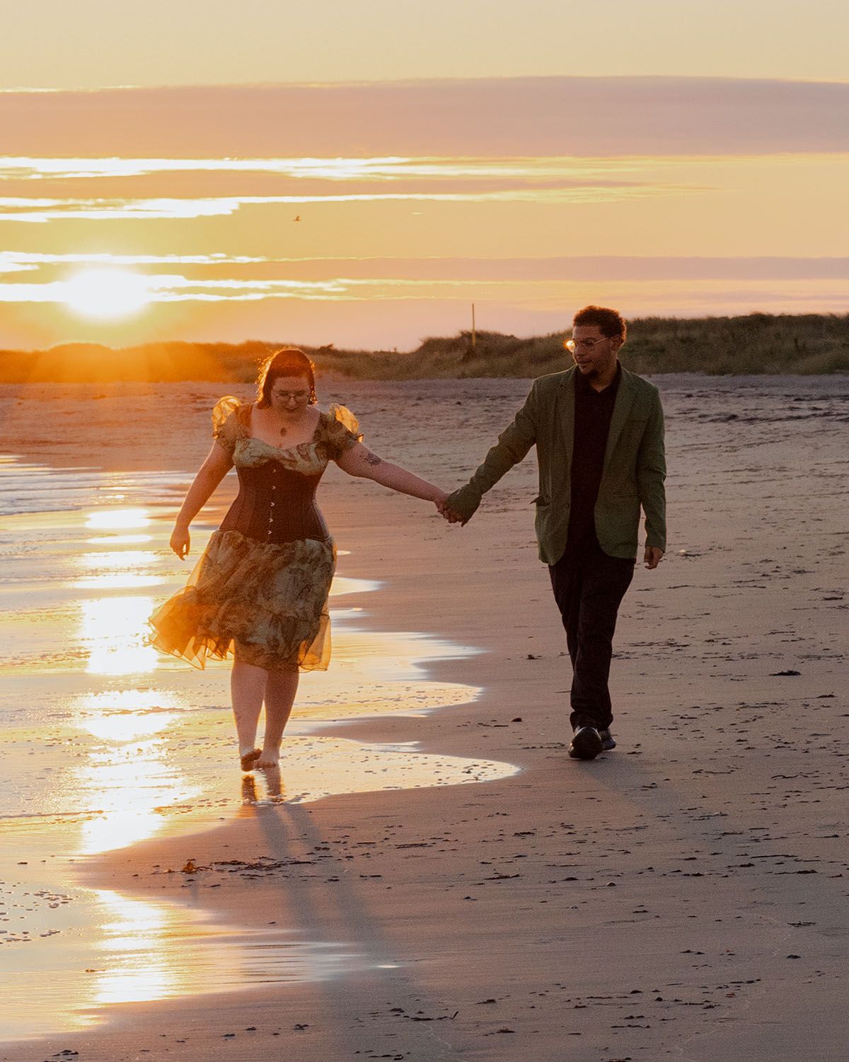 Two people walk hand in hand on a beach during a vibrant orange sunset.