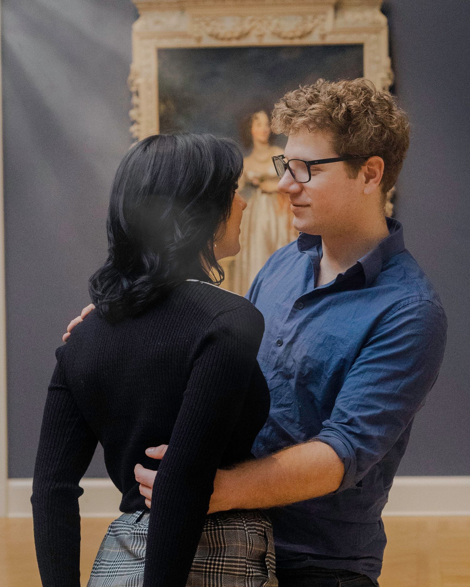 A young couple embraces romantically in an art museum gallery with a classical portrait painting in the background.