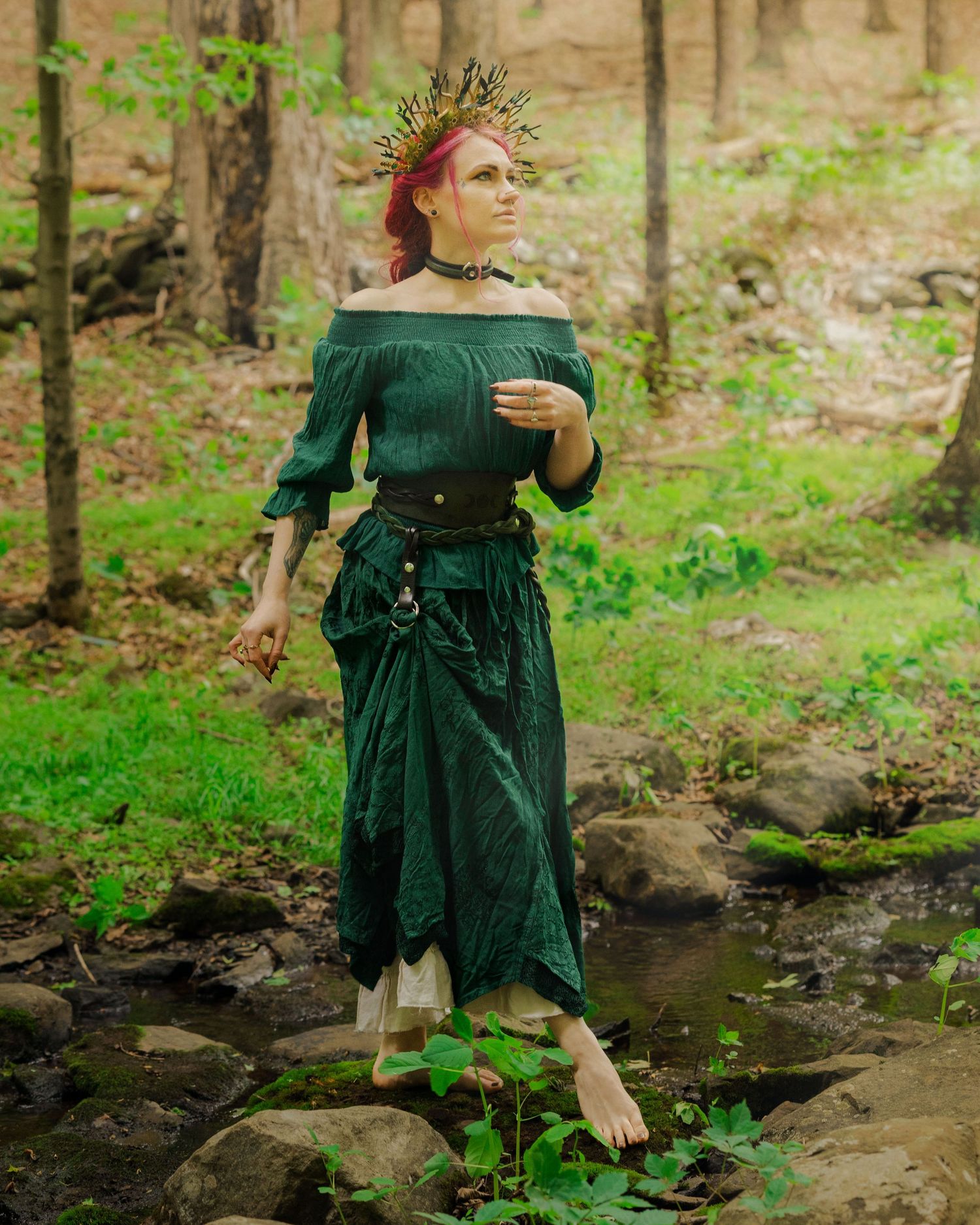Woman in green medieval dress and gold crown poses barefoot by a forest stream.