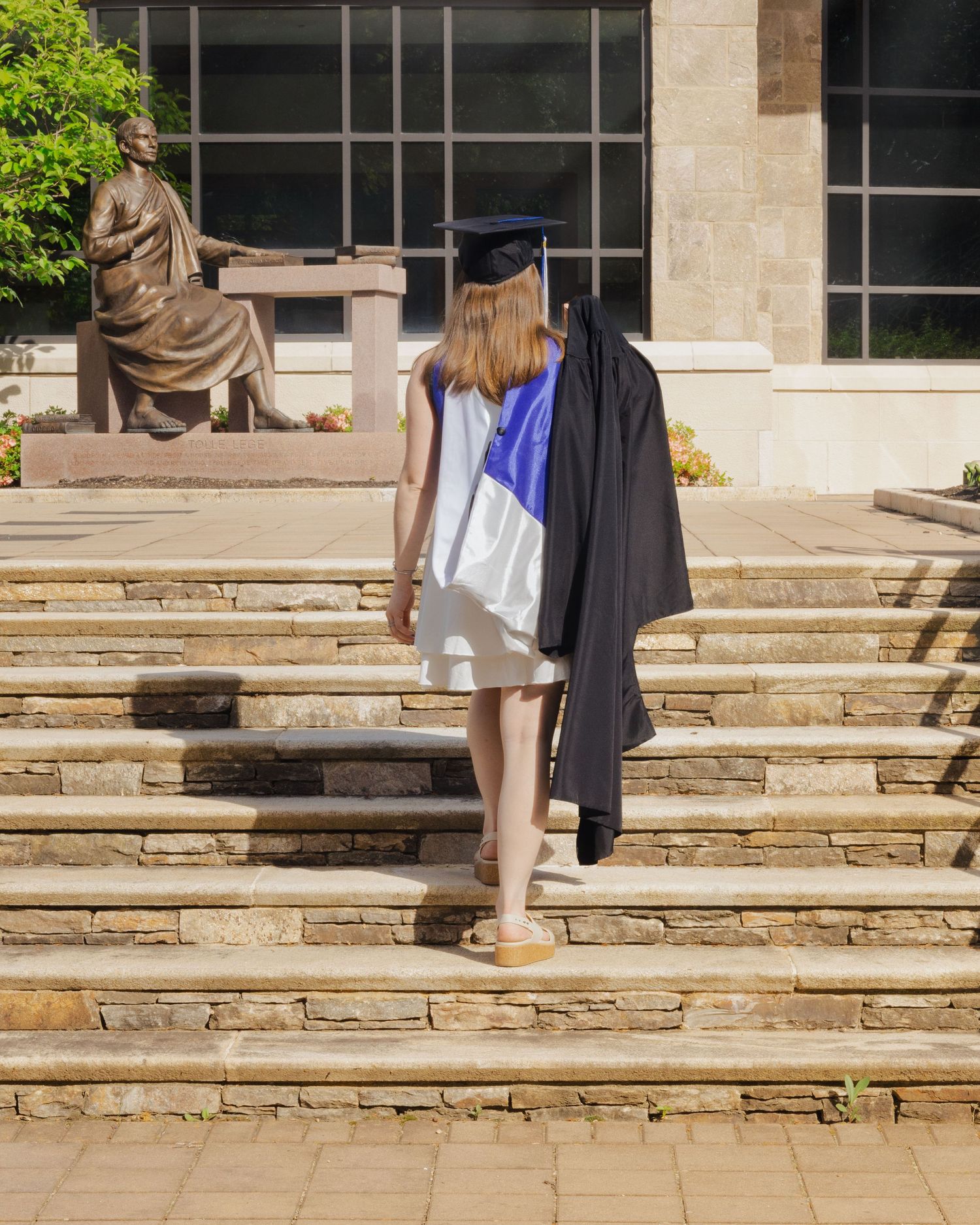 A graduate in cap and gown walks up stone steps toward a university building with a seated statue nearby.