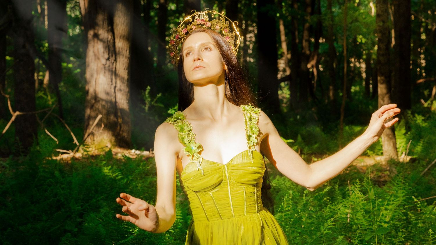A woman in a yellow dress and floral crown poses gracefully in a lush green forest setting.