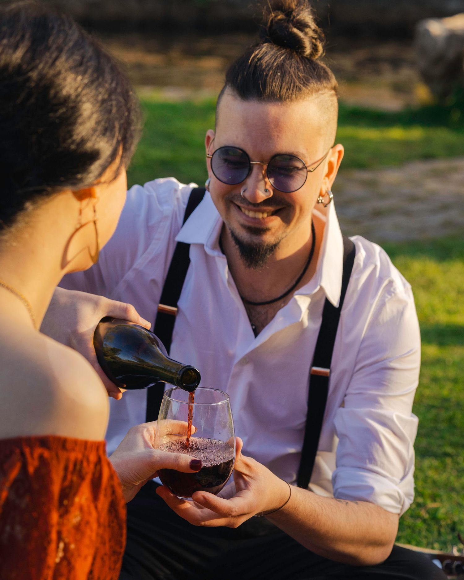 Smiling man with bun hairstyle and suspenders pouring red wine into a glass outdoors at sunset.