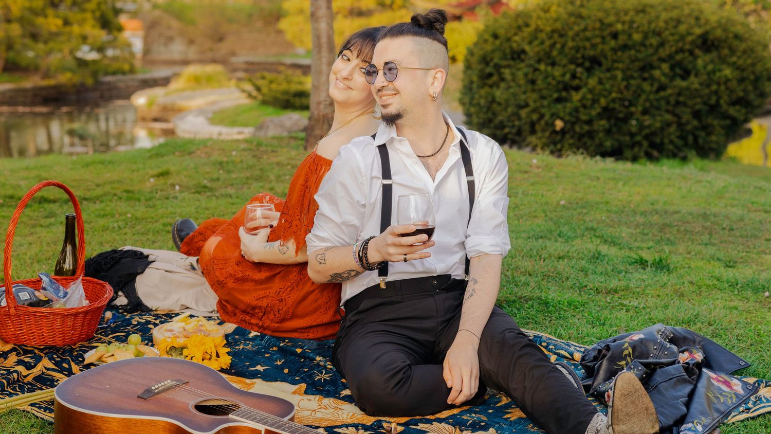Couple enjoying a romantic picnic in a park, sitting on a blanket with wine glasses and a guitar nearby.