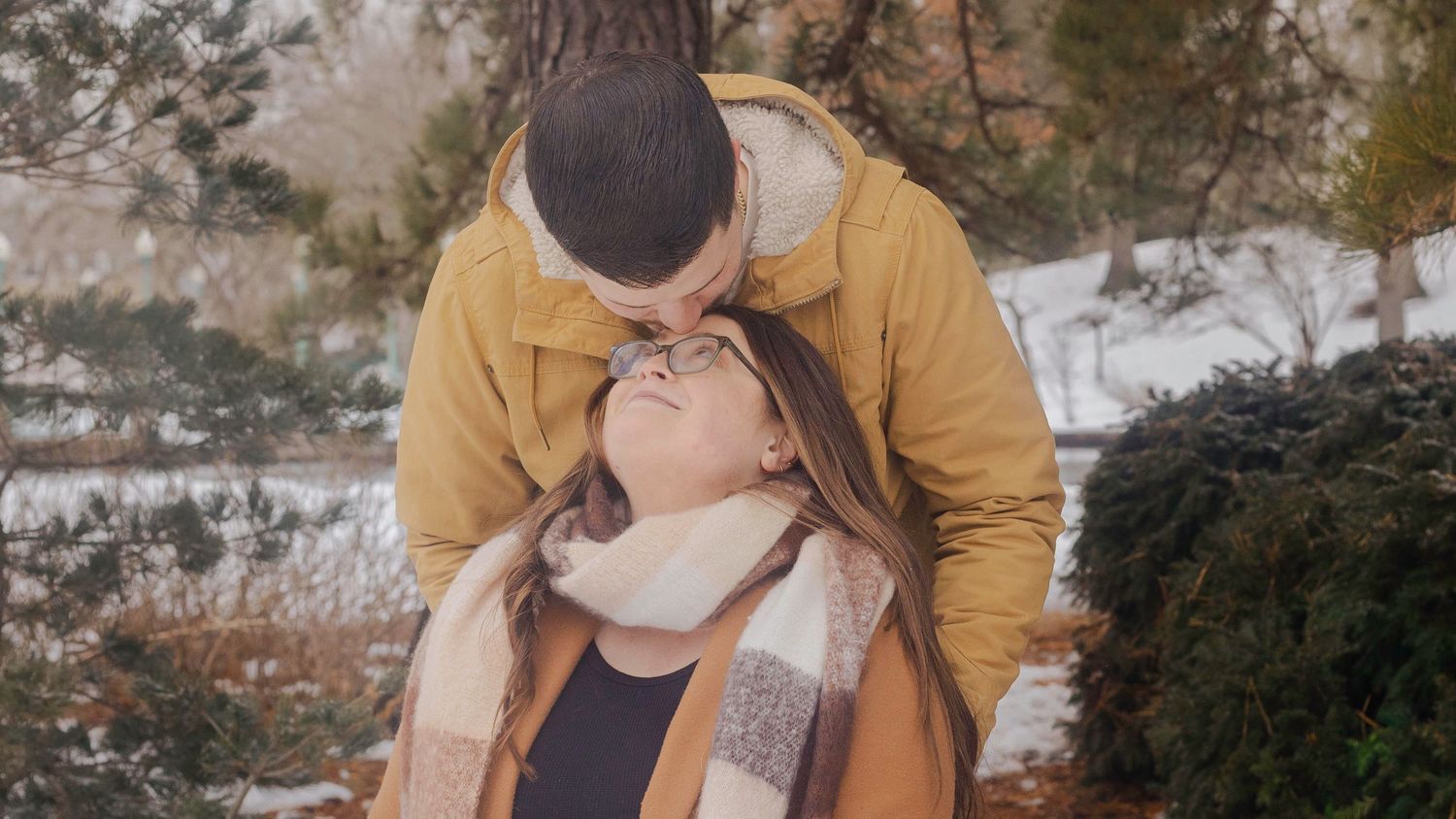 Couple sharing a tender moment outdoors in winter, man kissing woman's forehead near snow-dusted evergreen trees.