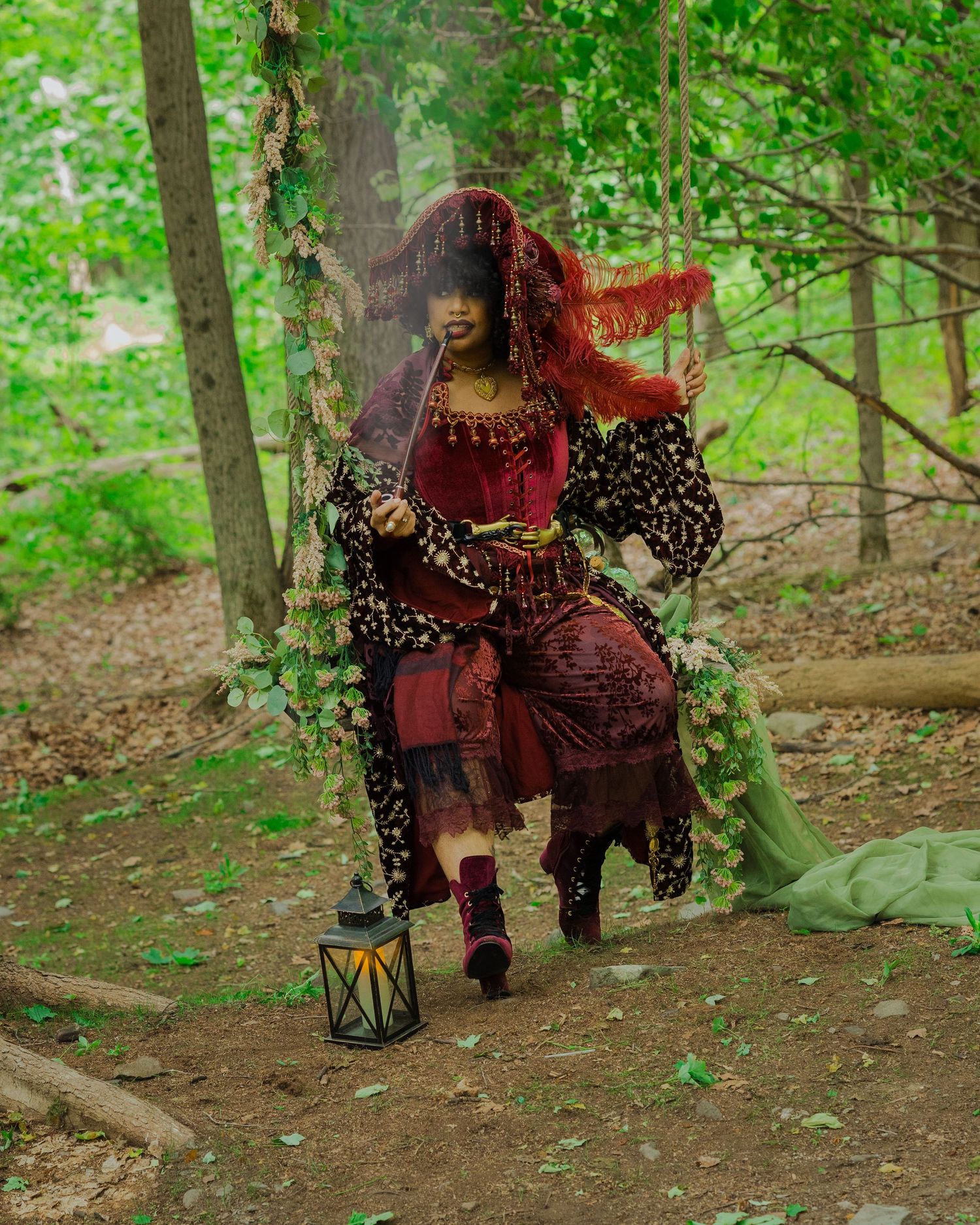 Woman in crimson Renaissance costume with veil sits on swing in enchanted forest setting with lantern.