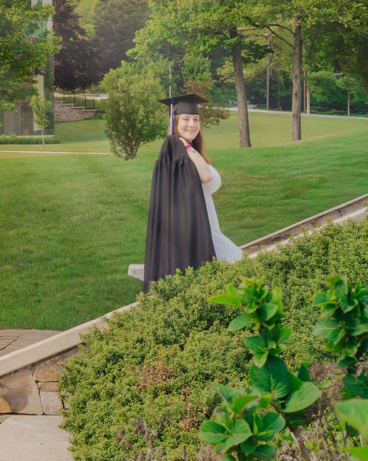 A smiling graduate in black cap and gown holds a diploma outdoors on a lush green campus lawn.