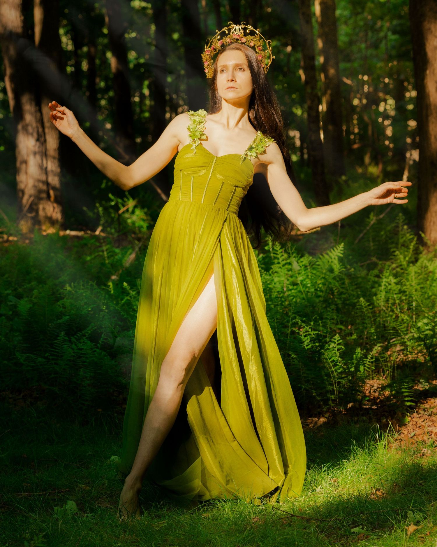 Woman wearing gold crown and olive chiffon gown stands elegantly in a lush green woodland setting.