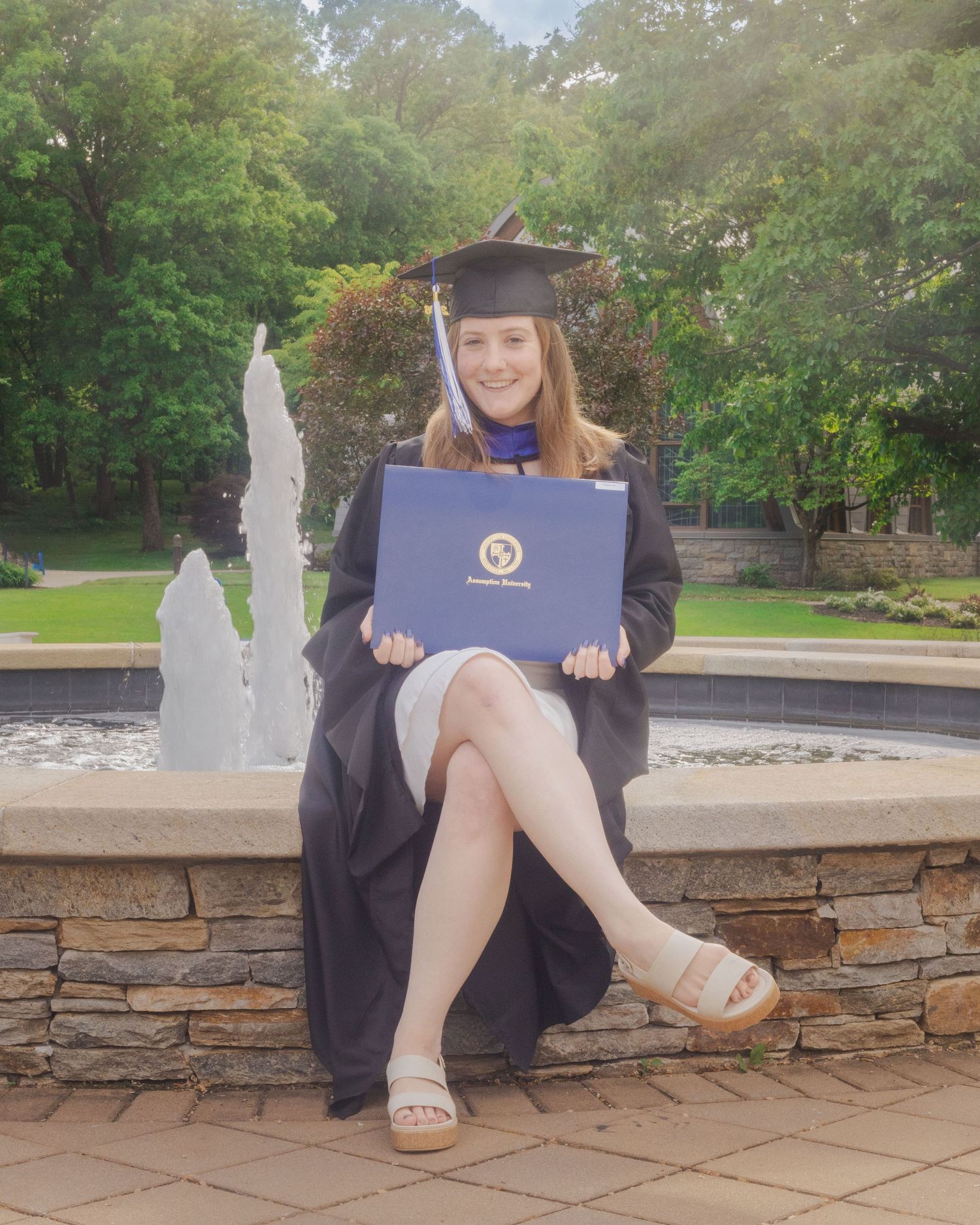 Young woman in graduation cap and gown holding diploma by fountain, celebrating academic achievement outdoors.