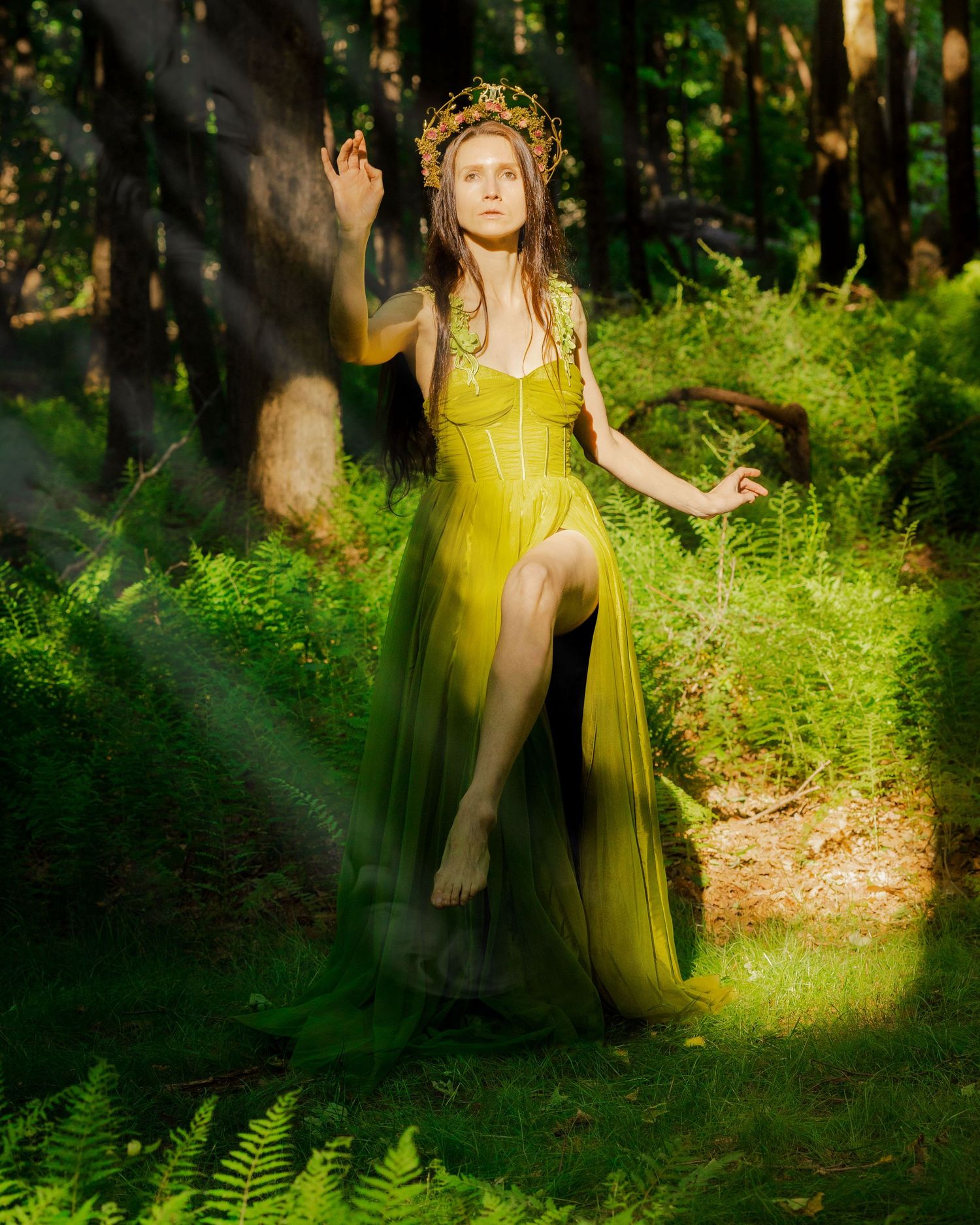 Woman in yellow-green gown and floral crown poses gracefully among ferns in a sunlit forest.