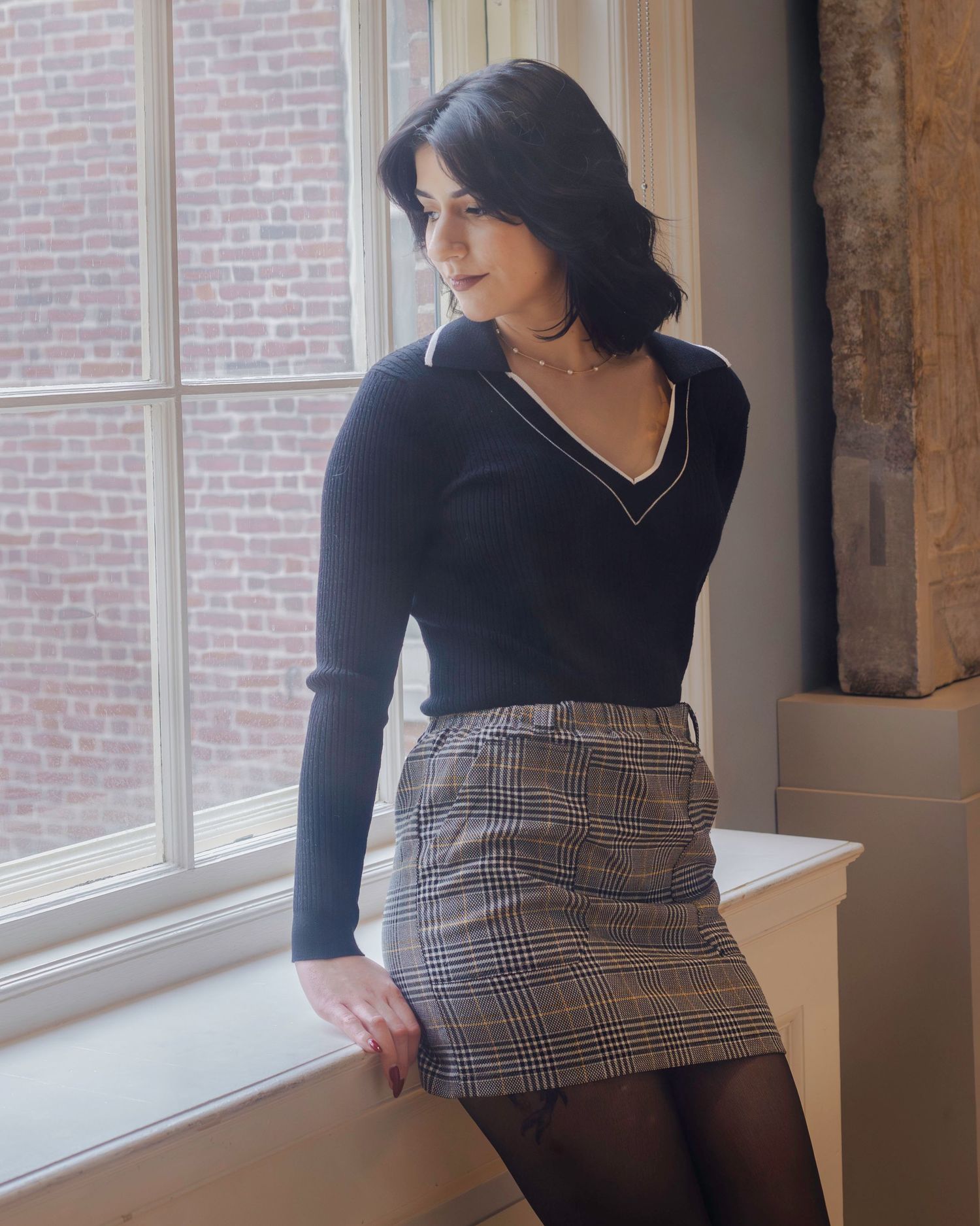 Woman in black V-neck sweater and plaid mini skirt standing by a window in a brick building.