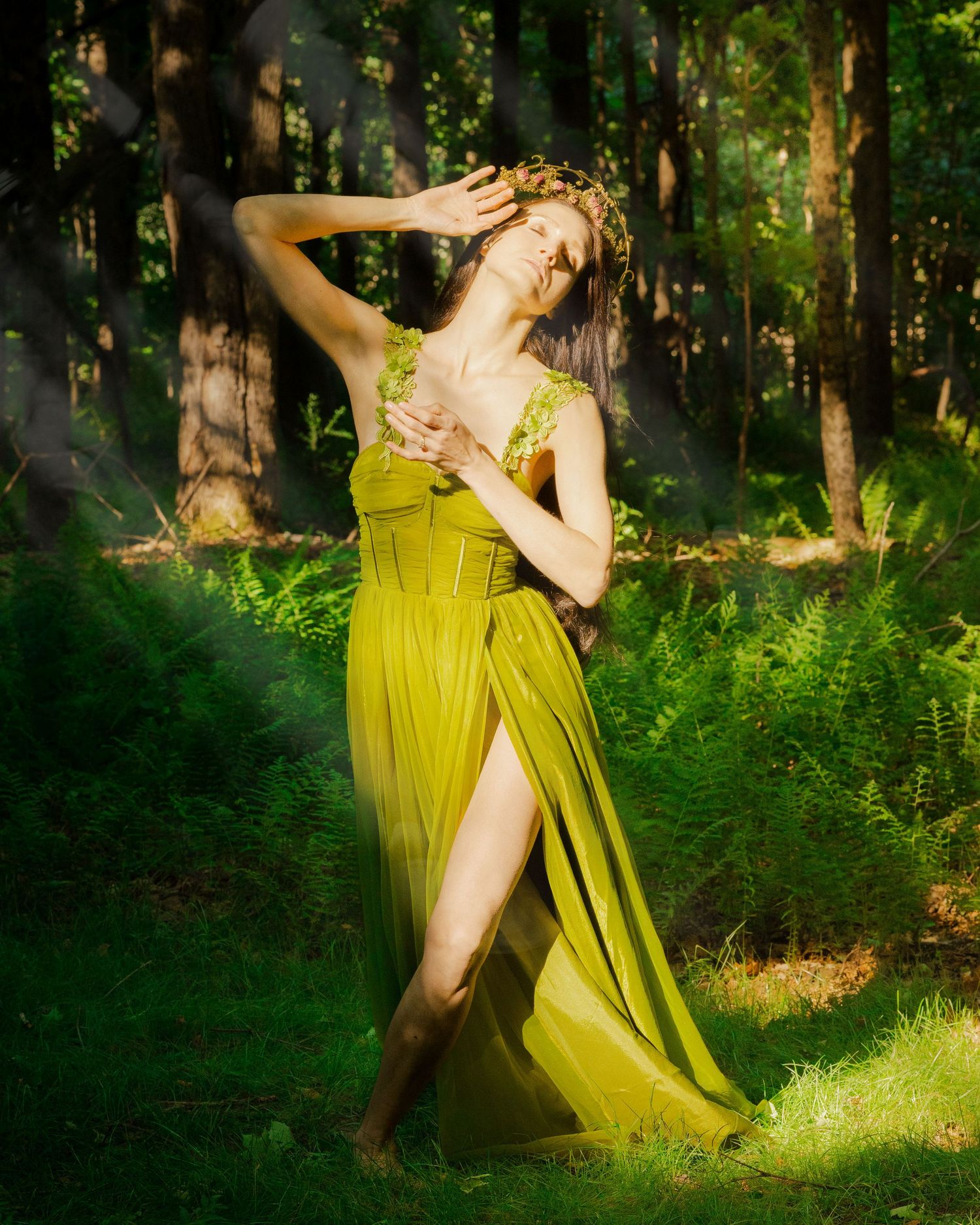 Woman in olive green gown with floral crown poses gracefully among ferns in a sunlit forest.