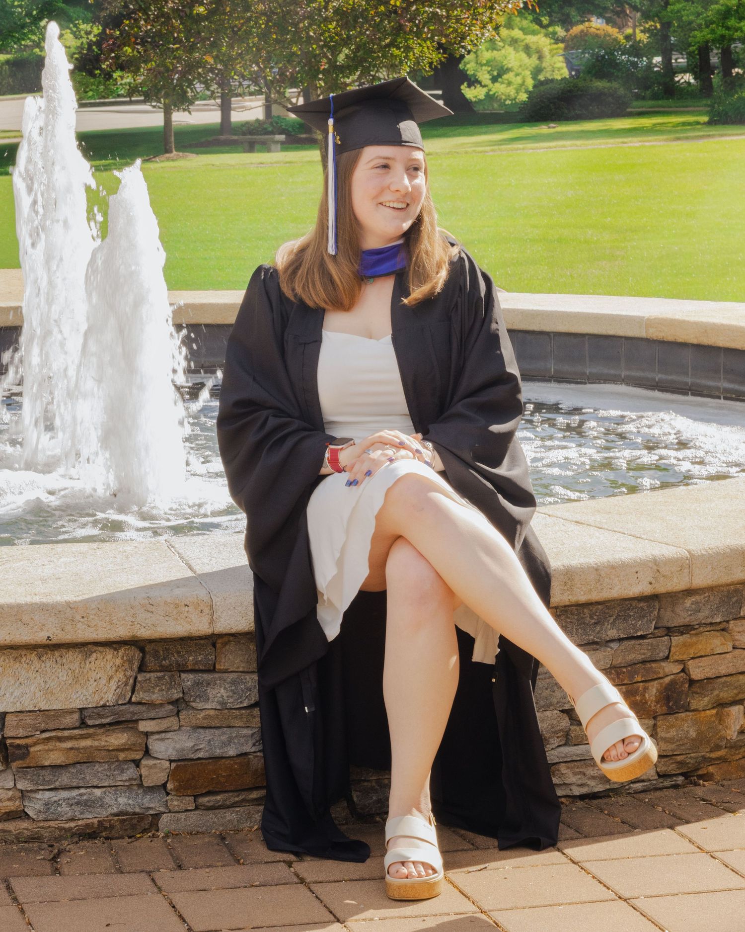 A smiling graduate in black cap and gown sits by a stone fountain on a sunny campus, wearing white dress and wedge sandals.