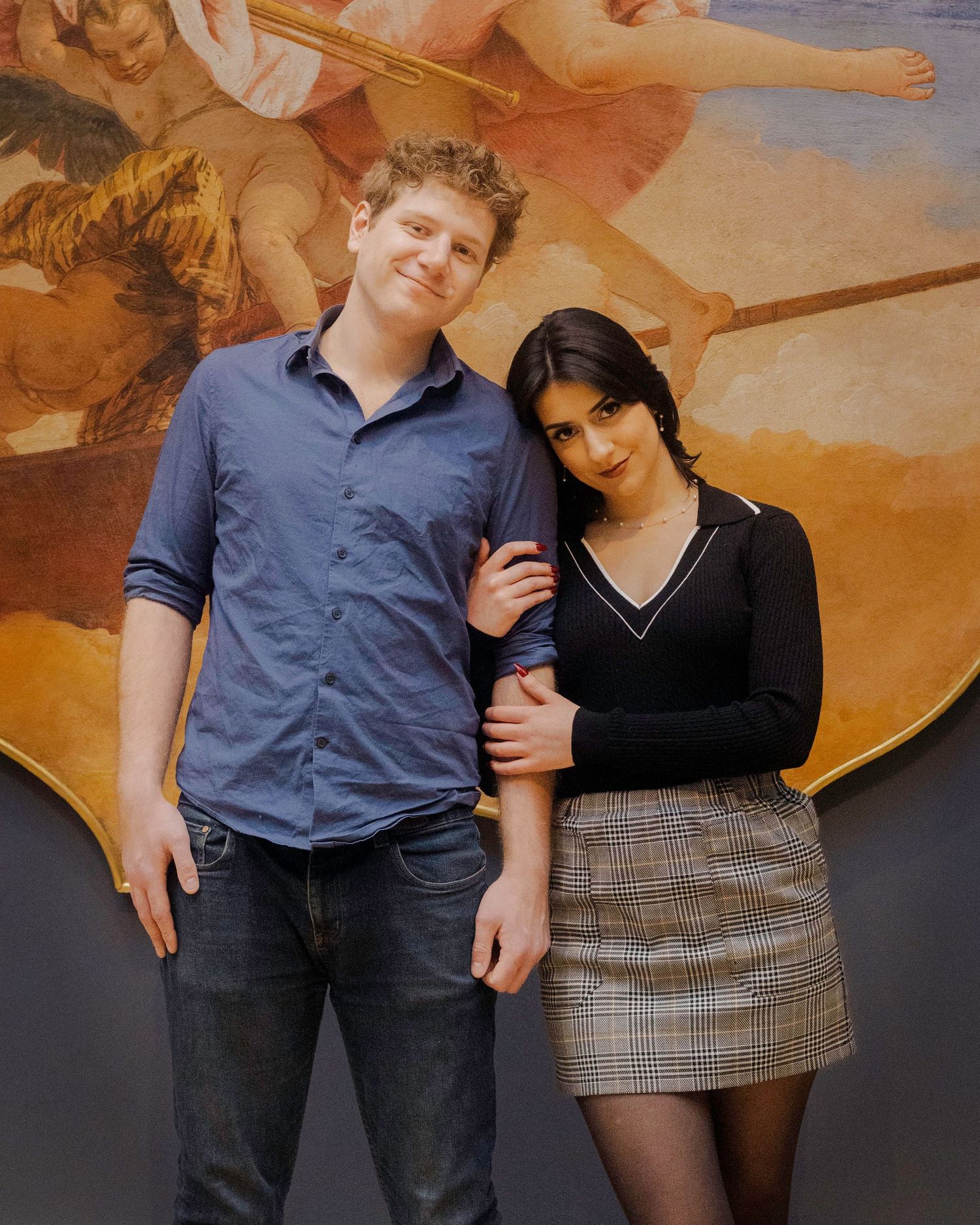 A young couple poses together in front of a classical Renaissance mural painting in an art gallery.