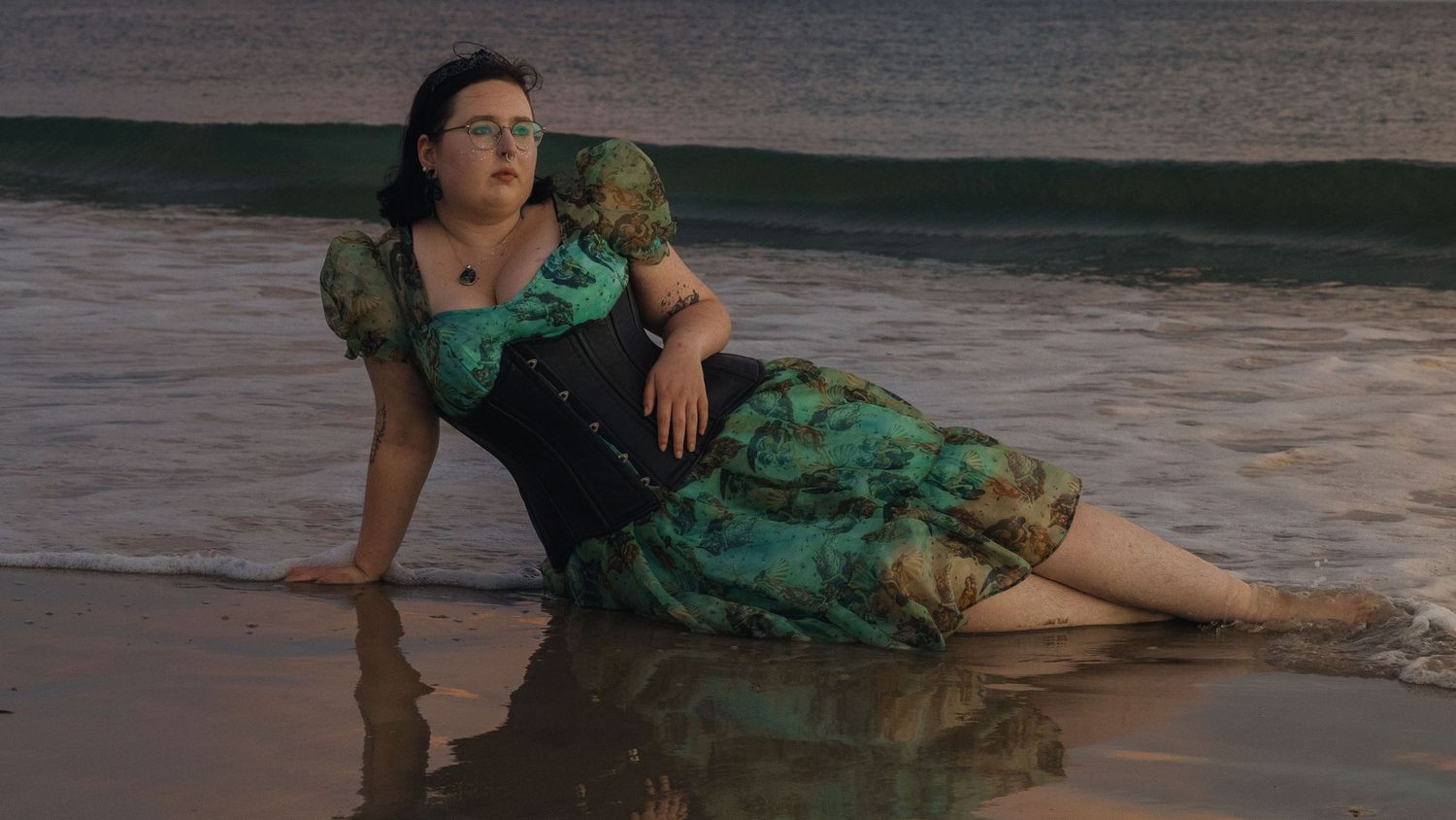 A person in a green floral dress poses dramatically on the wet sand at the beach during sunset.