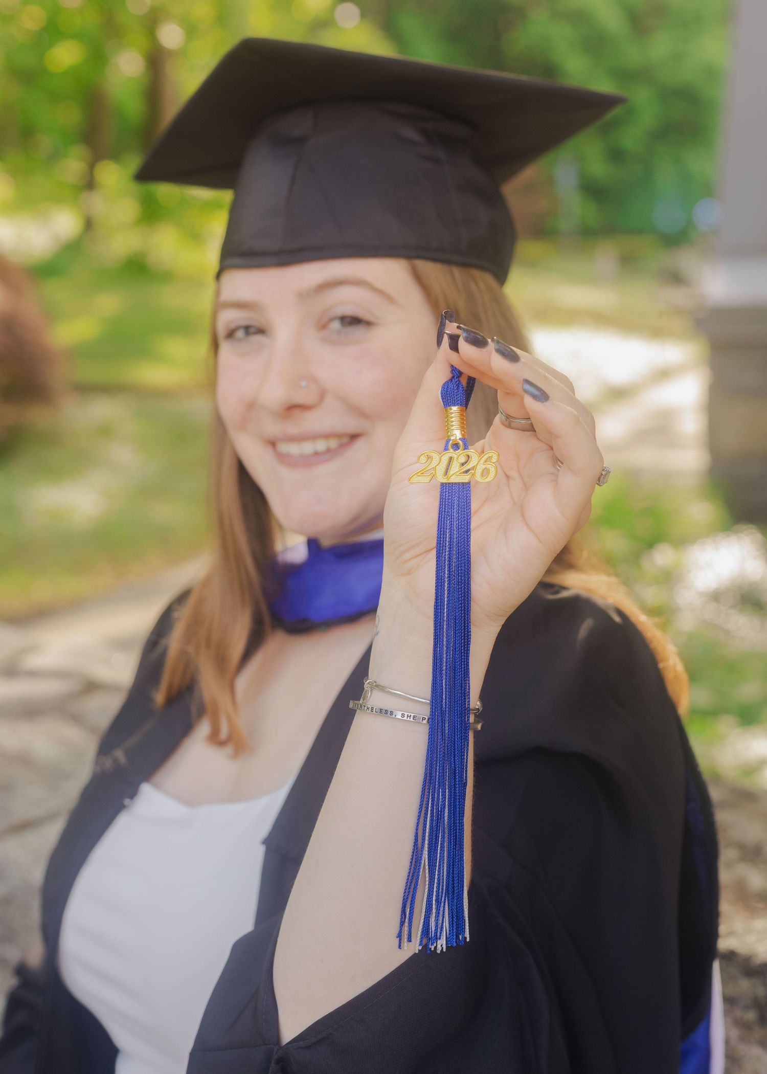 Smiling graduate in black cap and gown holds blue tassel with 2026 charm outdoors.