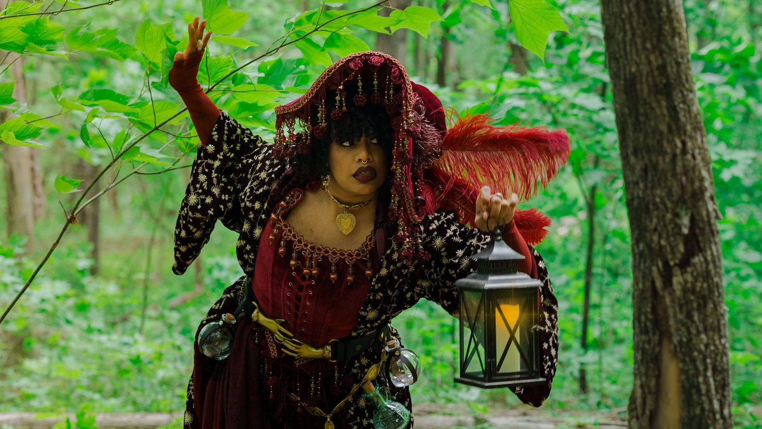 Woman in ornate red and gold costume holding lantern in forest setting.