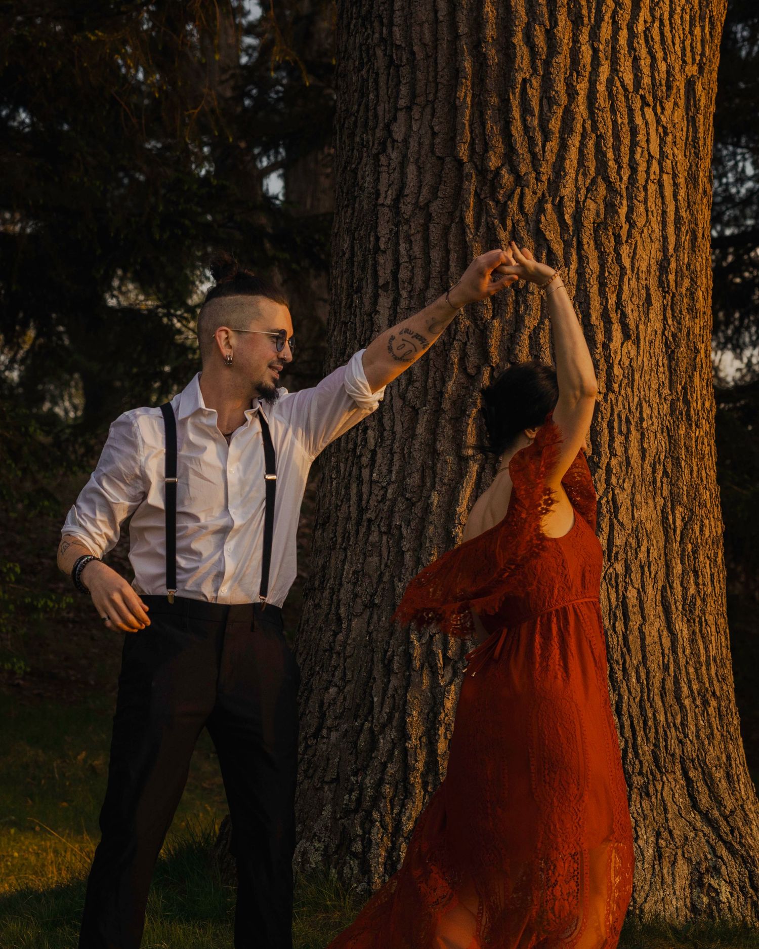 Couple dancing together near a large tree trunk in warm golden sunset light, woman in red dress.
