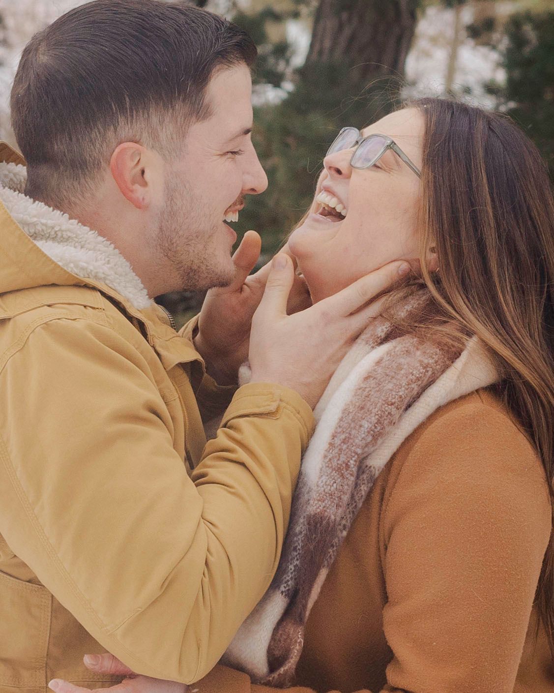 A couple in matching tan jackets laugh and share a tender moment outdoors in a snowy winter setting.