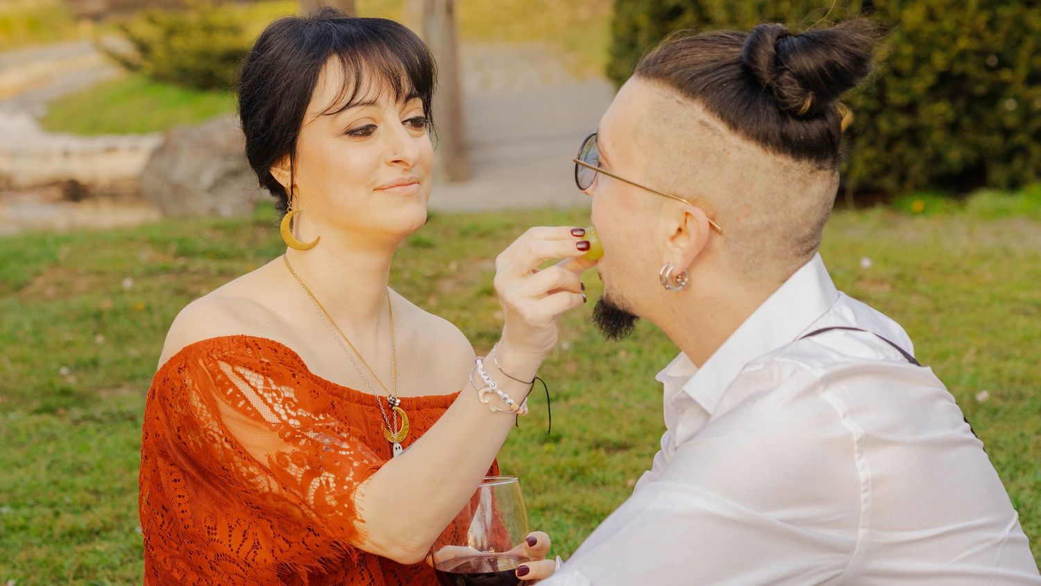 A couple shares a romantic moment outdoors, woman in red lace top feeding her partner a small flower in a green park.