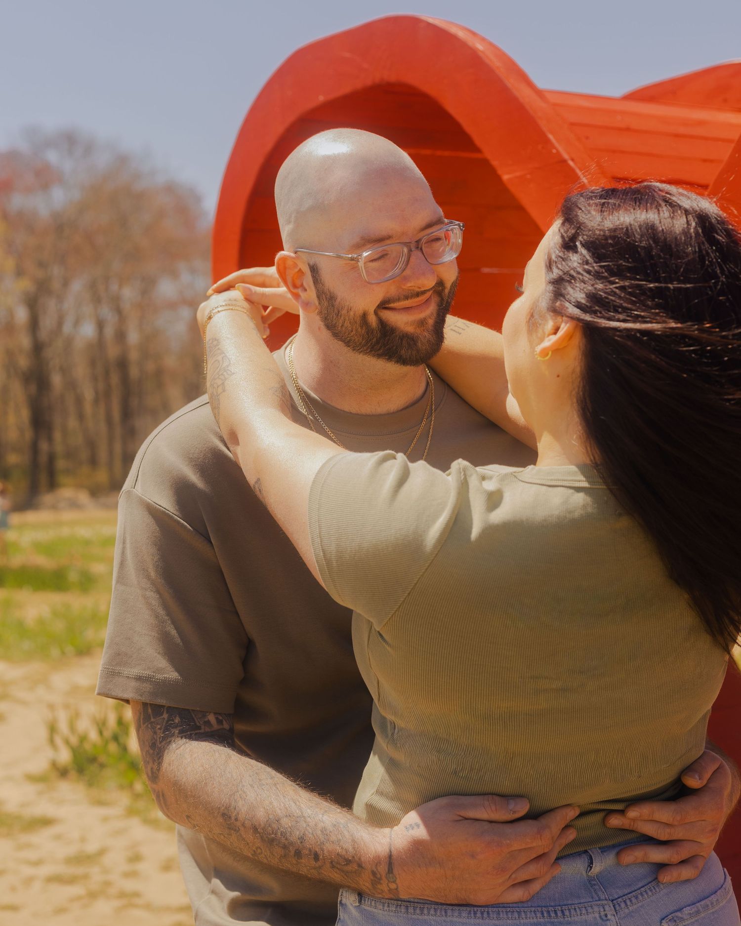 A couple embraces outdoors near a red heart sculpture on a sunny day, smiling at each other.