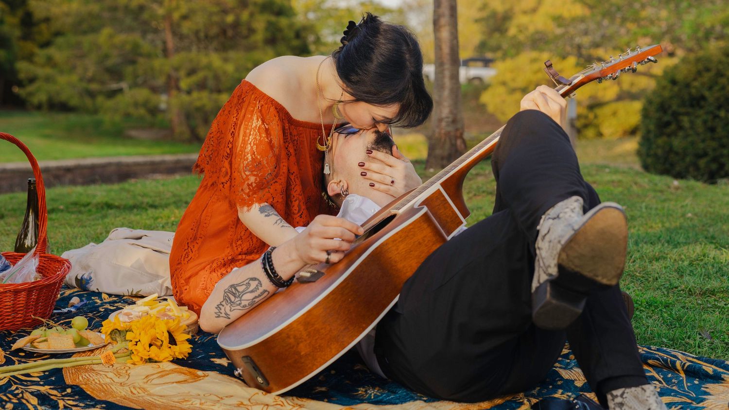 Woman in orange dress playing guitar with partner during romantic outdoor picnic on green grass in park.