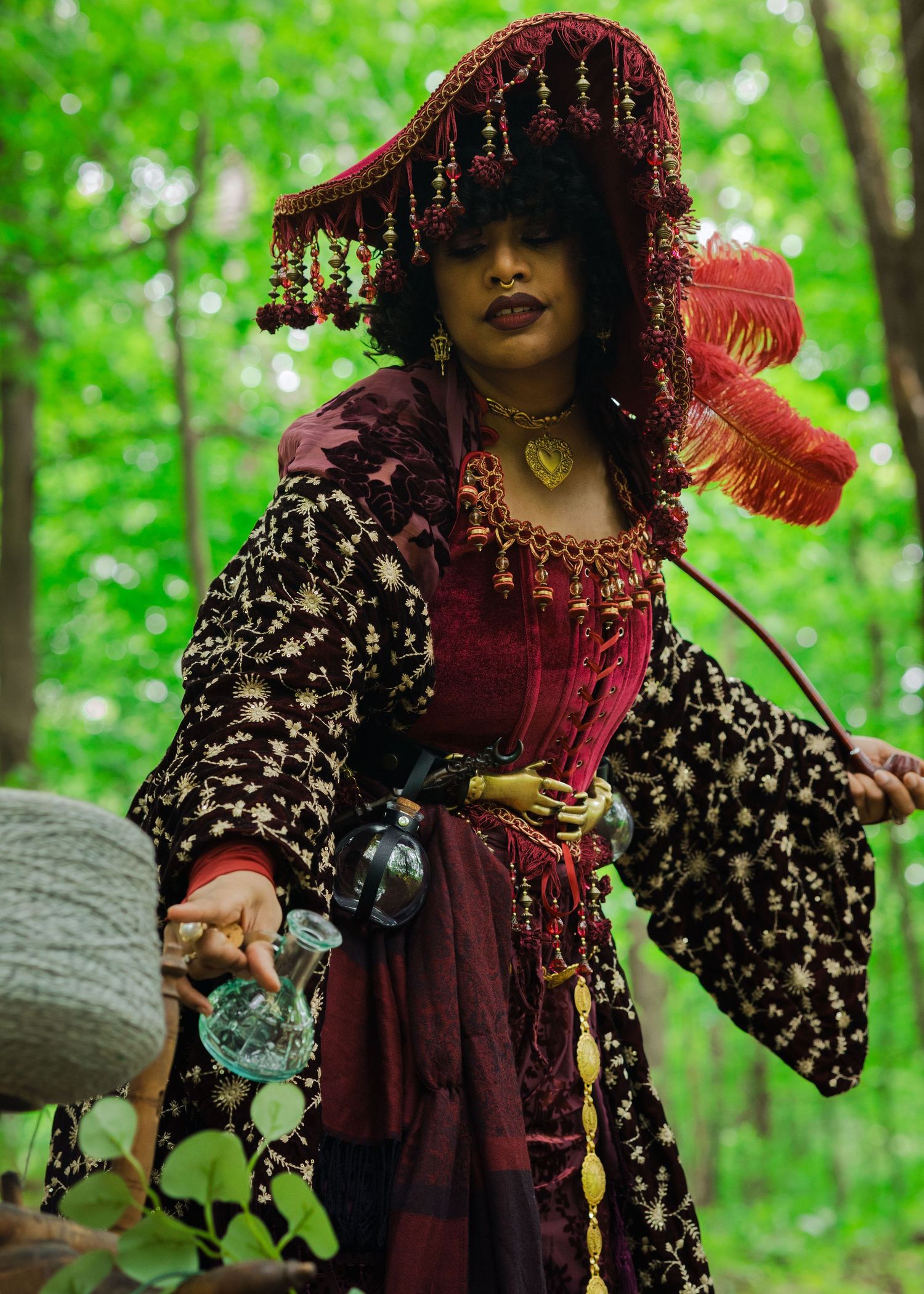 Woman in elaborate burgundy costume with ornate headdress and feathers poses in forest setting.