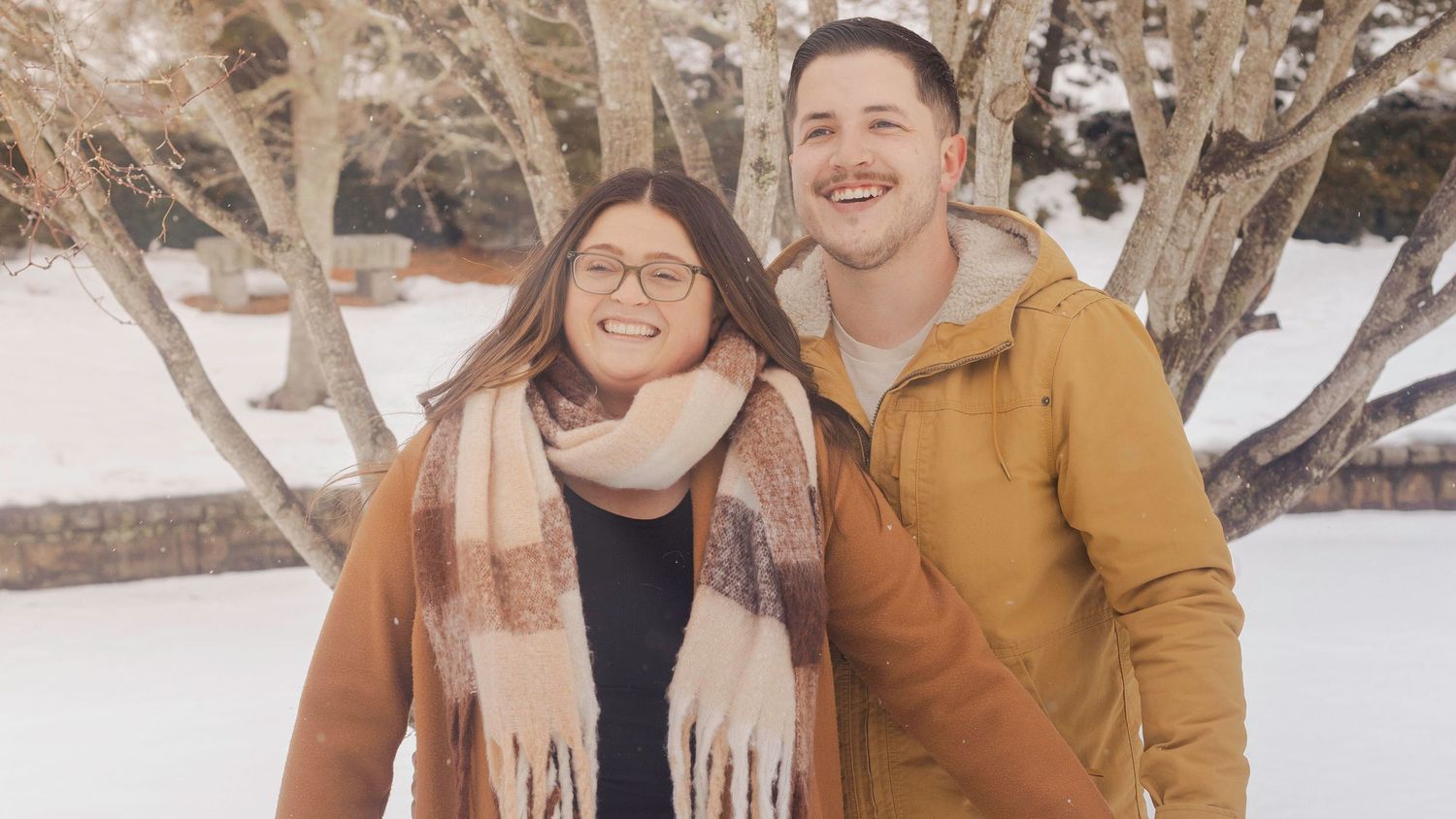 Couple smiling together outdoors in winter, woman in plaid scarf and brown coat, man in mustard jacket, snow-covered trees behind.